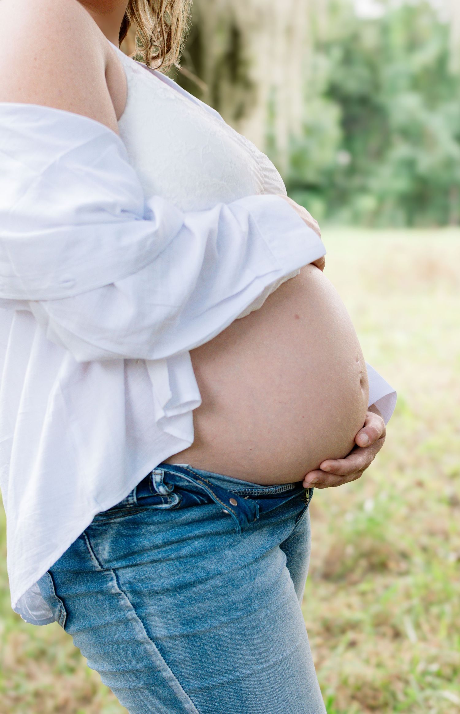 Pregnant woman in white shirt and jeans cradling her bare baby bump outdoors in a natural setting.