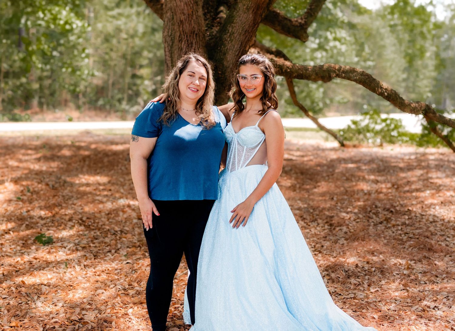 Two women posing outdoors under a large tree, one in a blue top and one in a light blue ball gown.