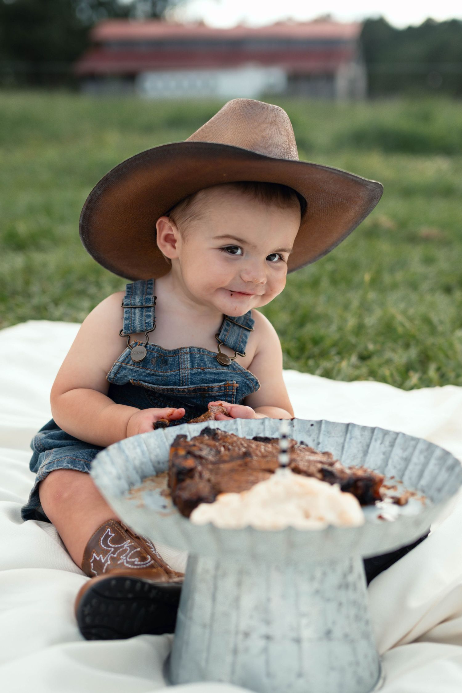 Smiling baby in cowboy hat and denim overalls sitting outdoors with chocolate cake on a white blanket.