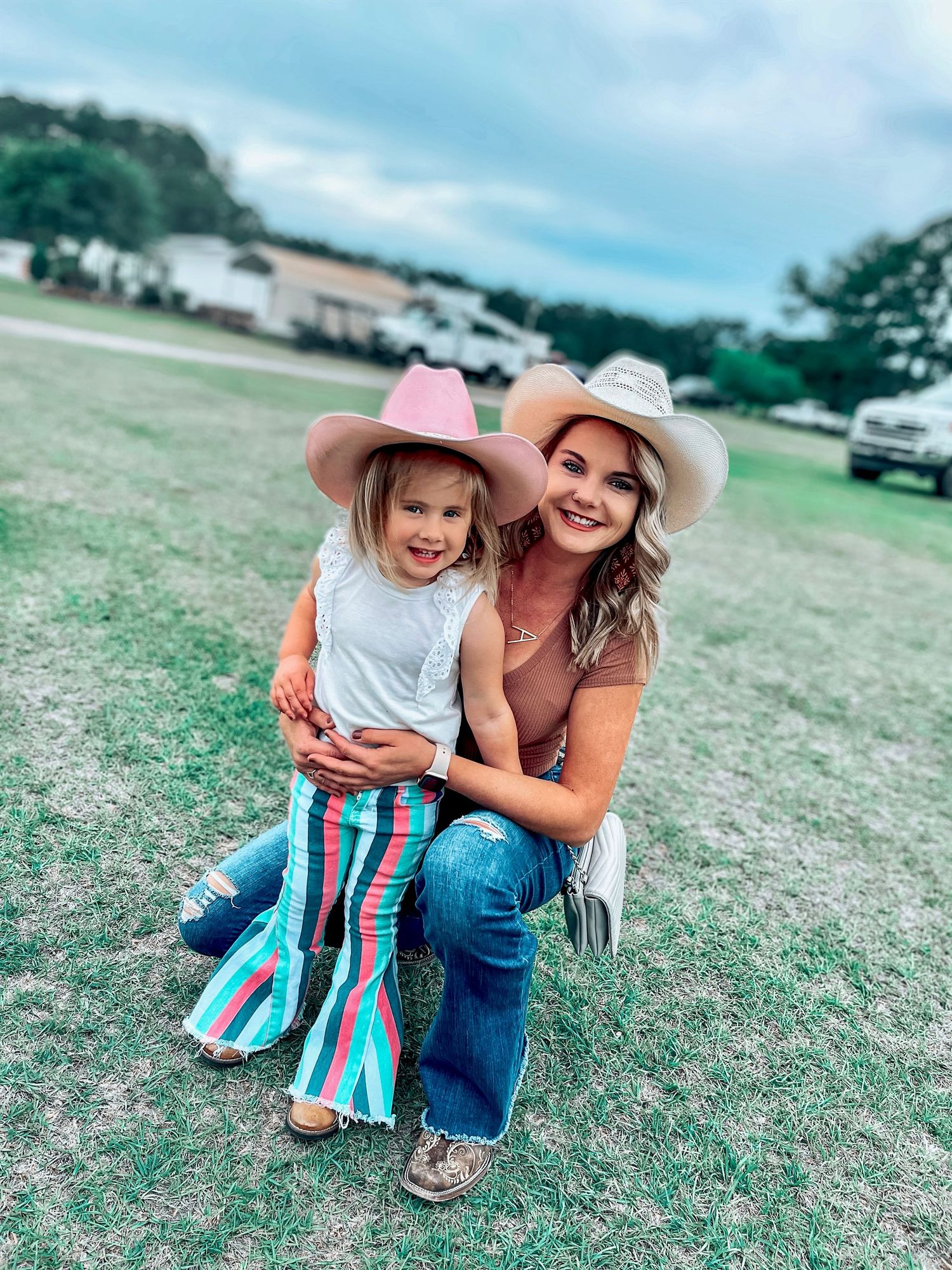 A woman and young girl wear matching pink cowboy hats and pose together on a grassy field at an outdoor event.