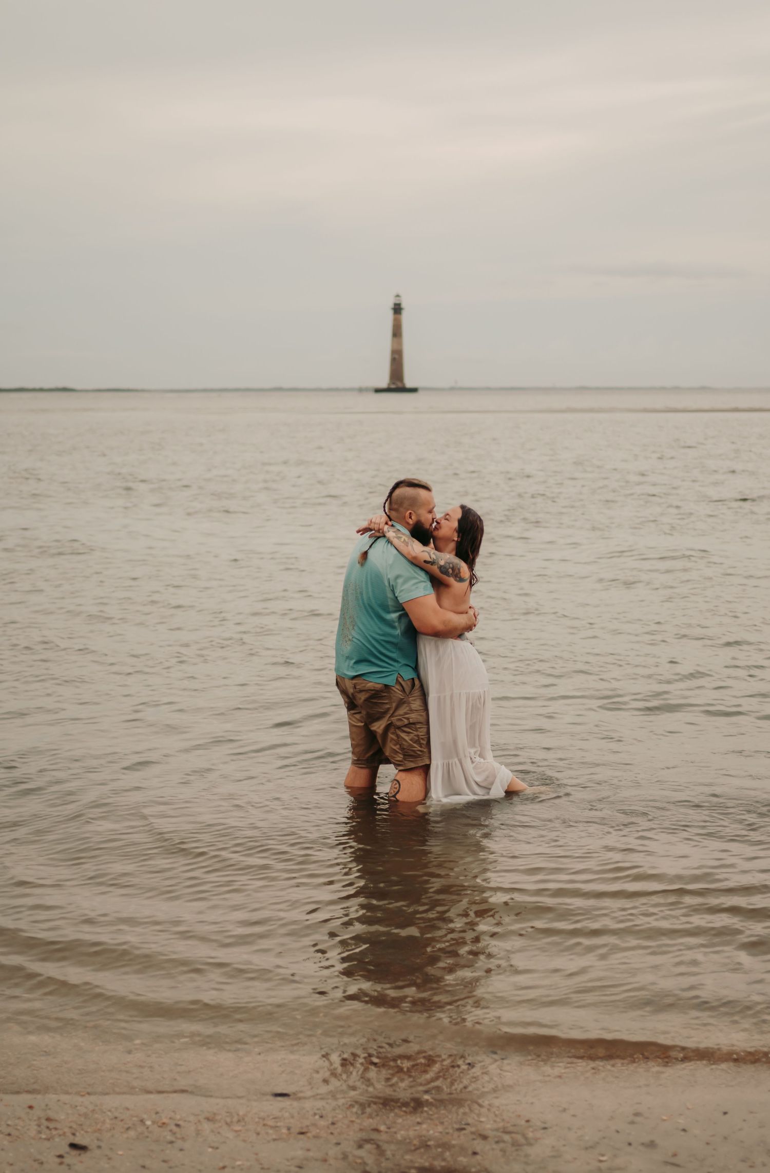 Couple kissing while standing in shallow ocean water with a lighthouse in the background on a cloudy day.