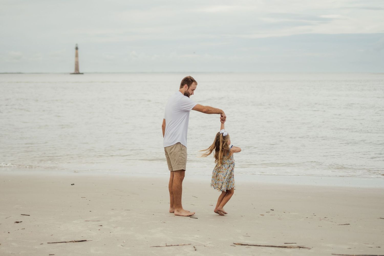 Father and daughter dancing on a sandy beach with a lighthouse in the background on a cloudy day.