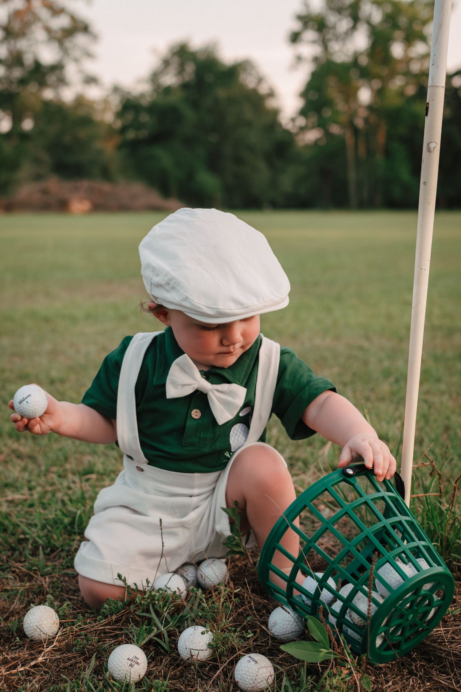 Adorable toddler in suspenders and flat cap collecting golf balls into a green basket on a golf course.