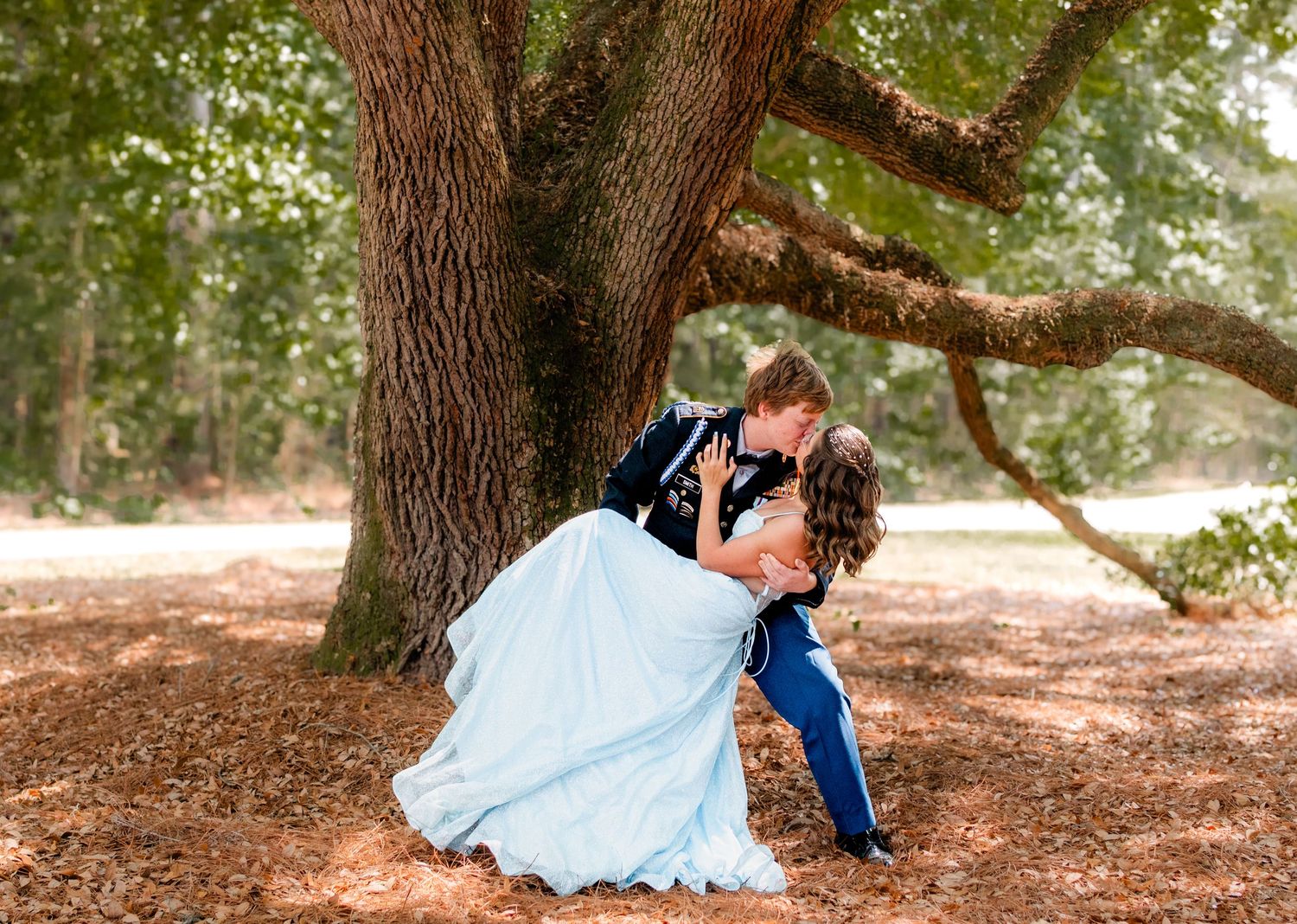 Couple sharing romantic dip kiss under large oak tree, man in military dress uniform.