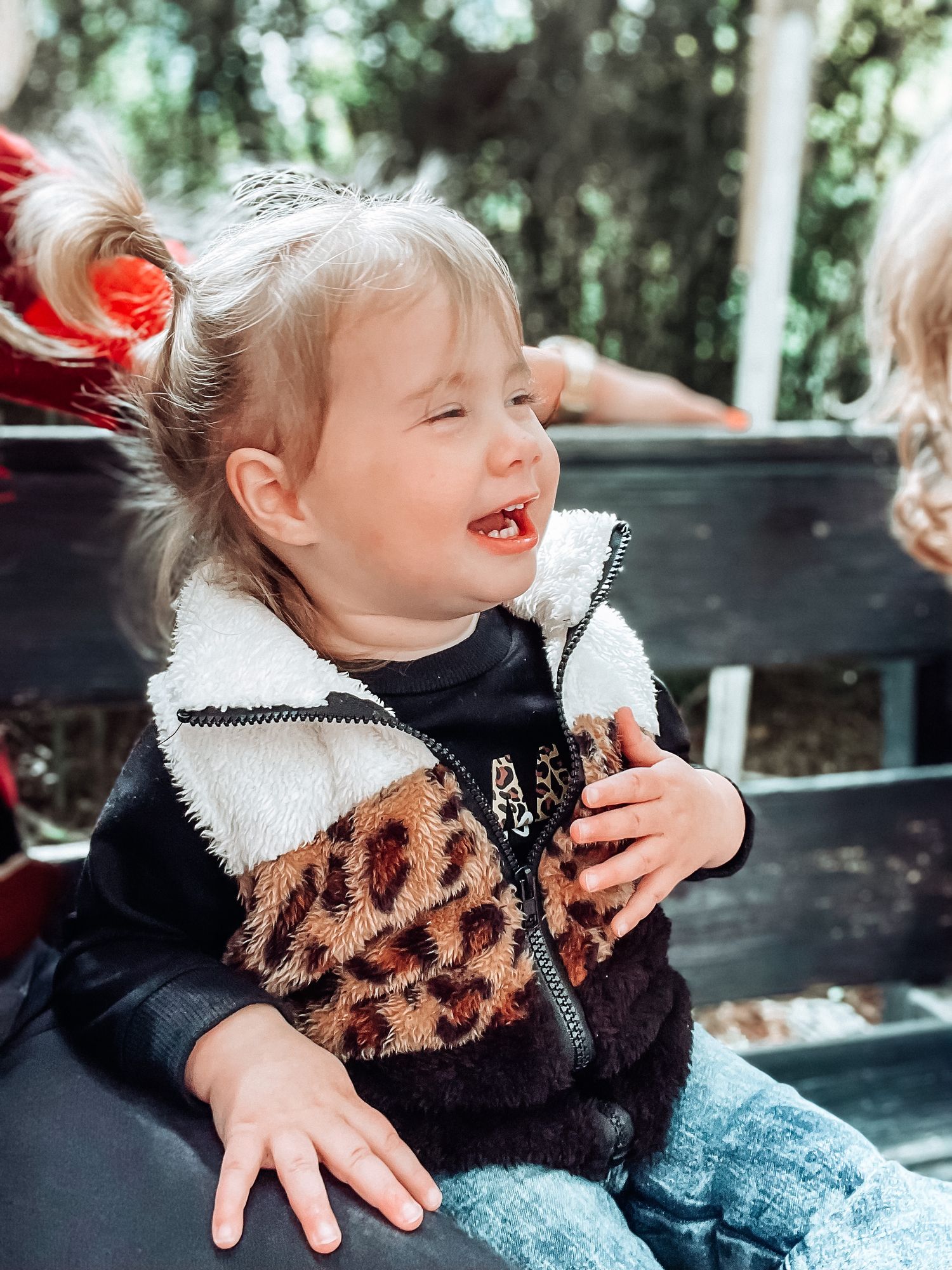 Laughing toddler girl in leopard print vest sitting on bench during outdoor adventure.