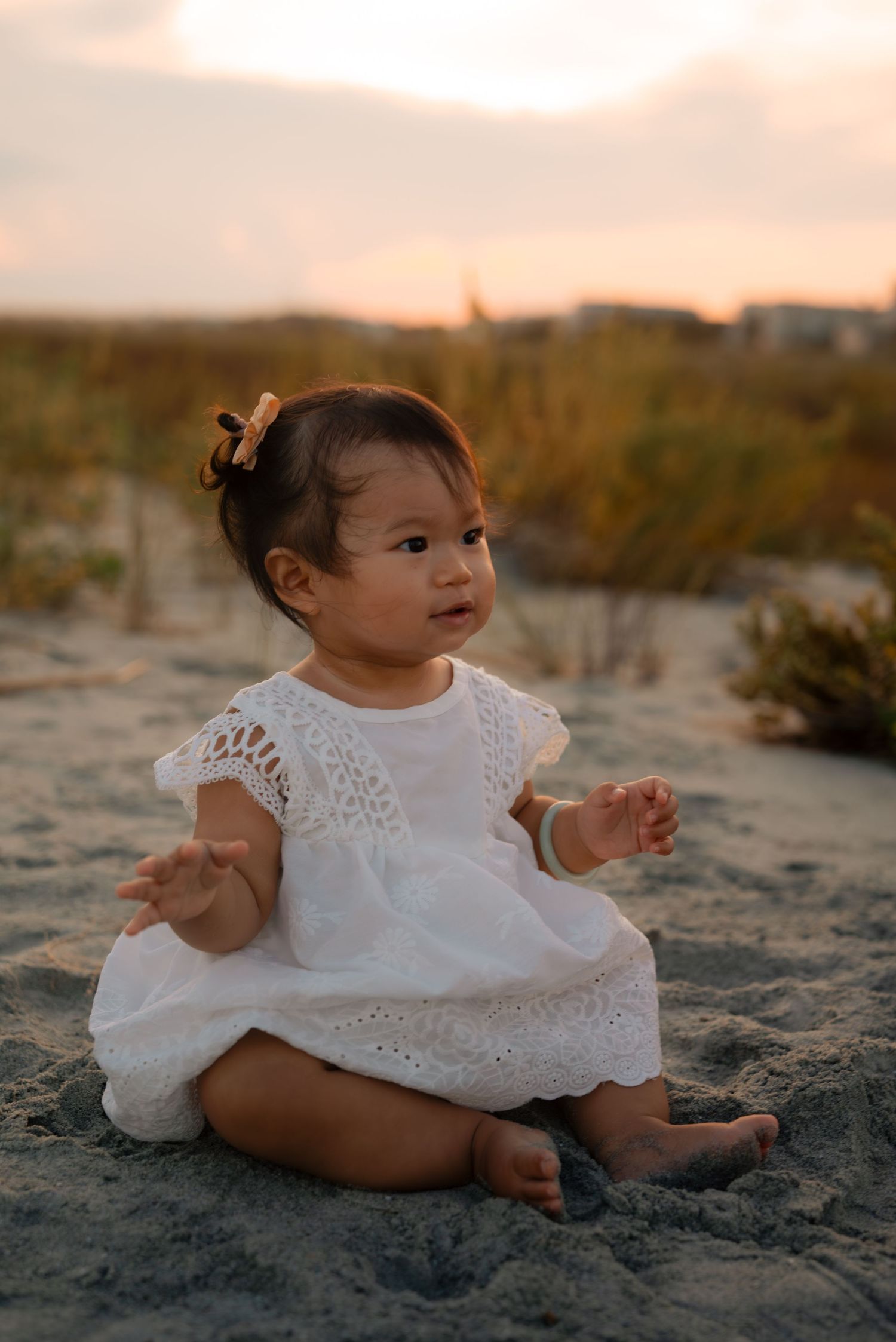A baby girl in a white lace dress sits on sandy beach at sunset, looking up curiously with a hair bow.
