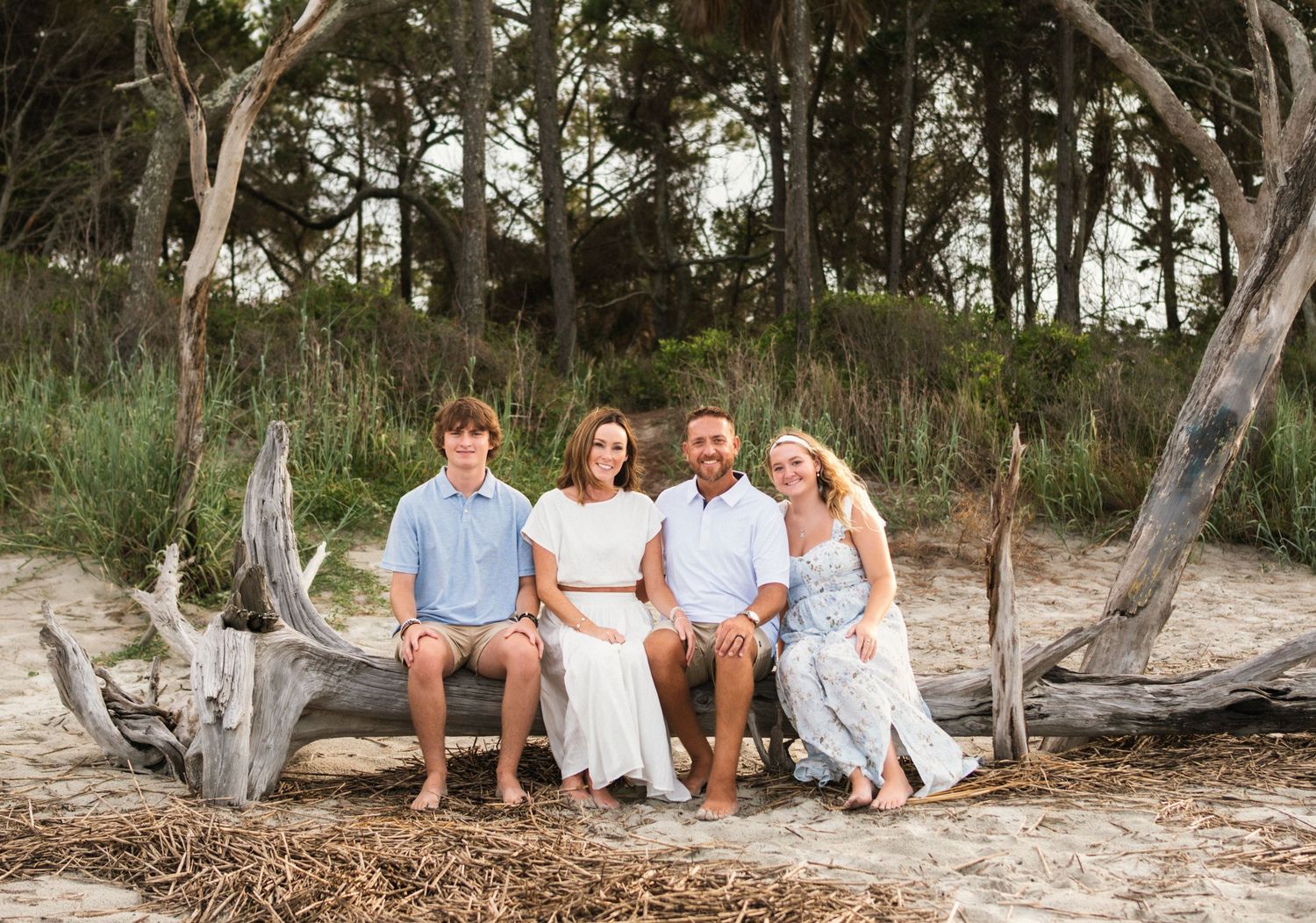Family of four smiling together on driftwood at a sandy beach with lush green trees in the background.