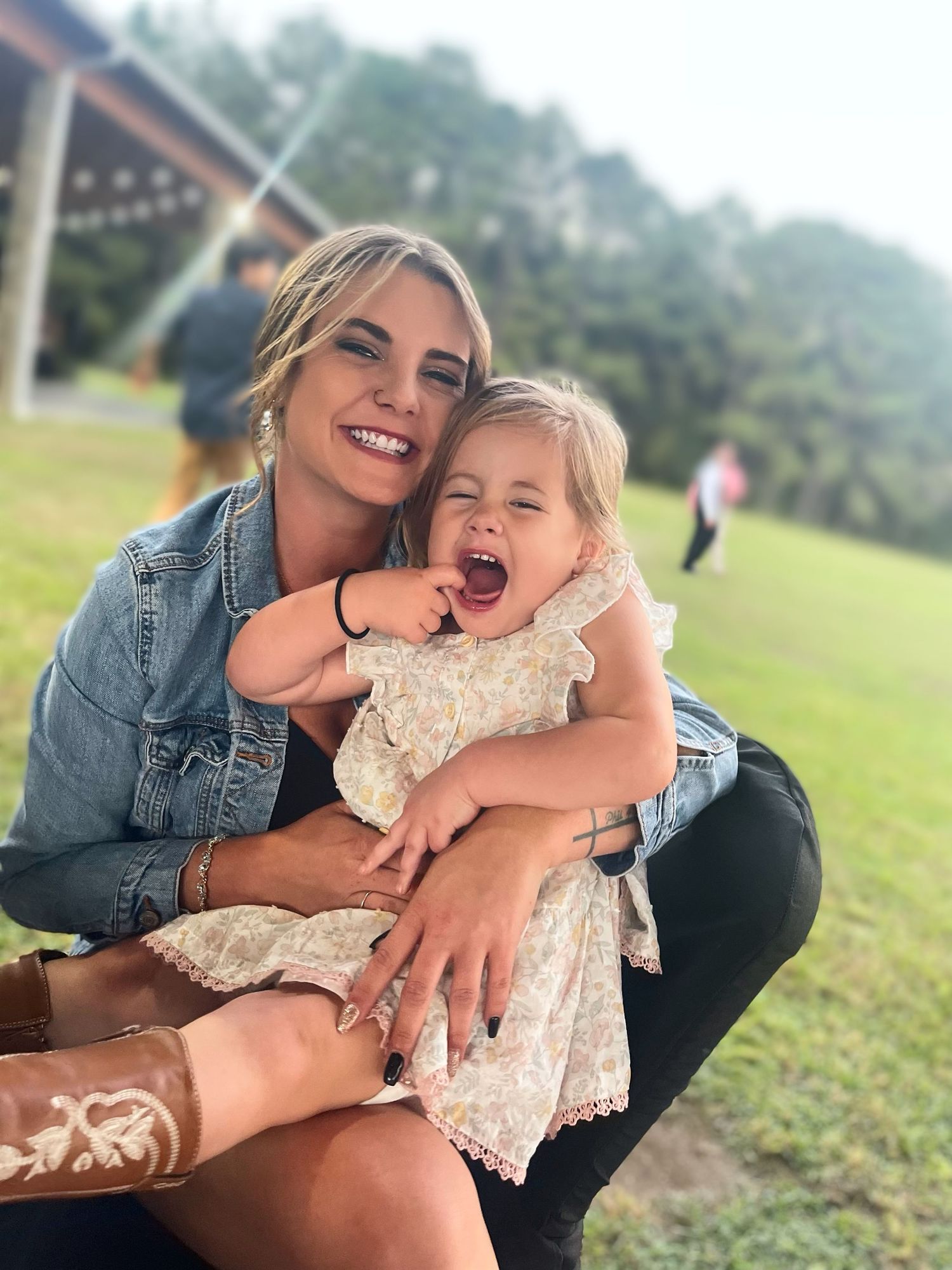 Happy woman in denim jacket hugging laughing toddler girl outdoors on green lawn.