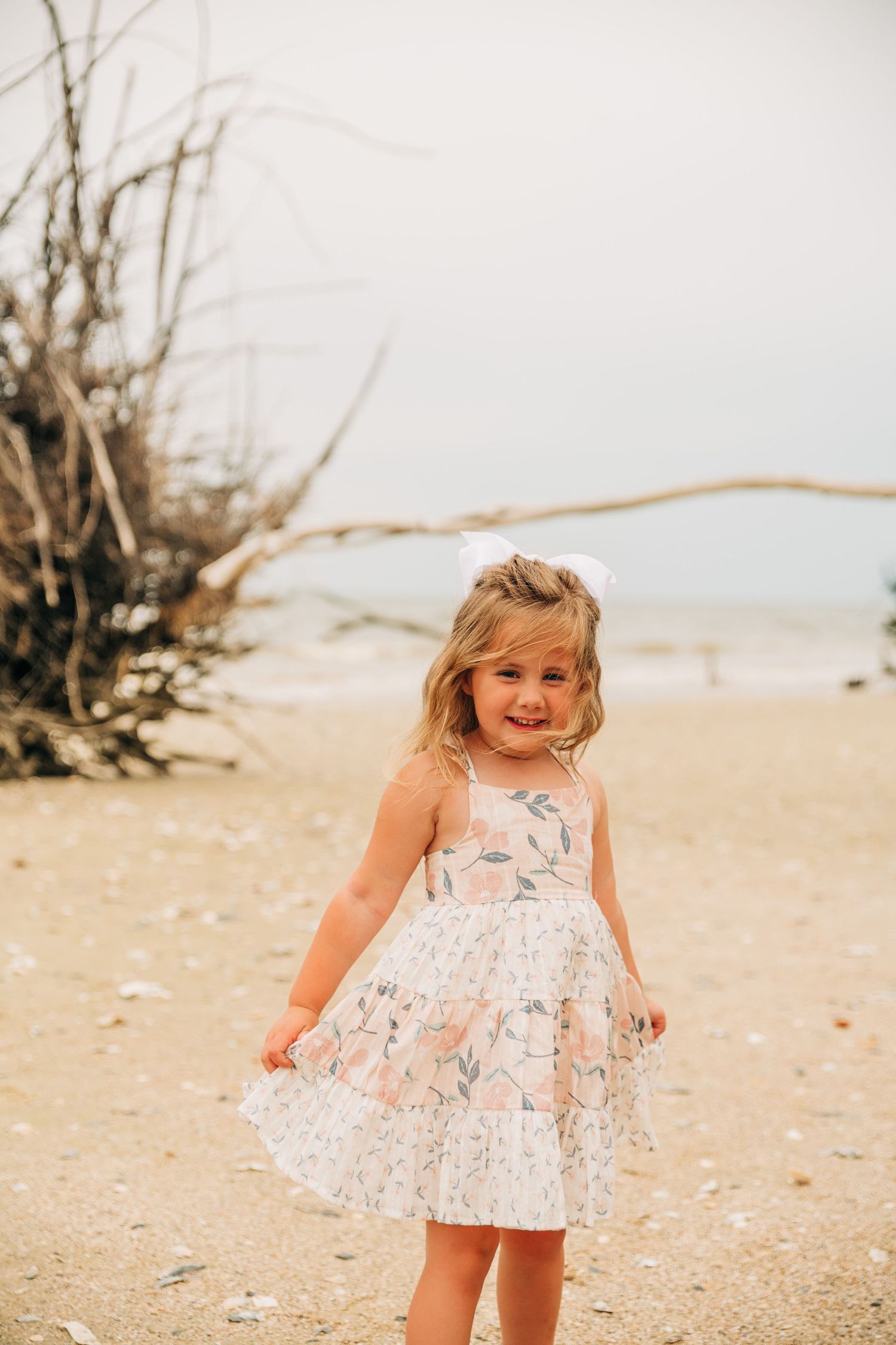A smiling little girl in a floral dress with a white bow twirls on a sandy beach with driftwood behind her.