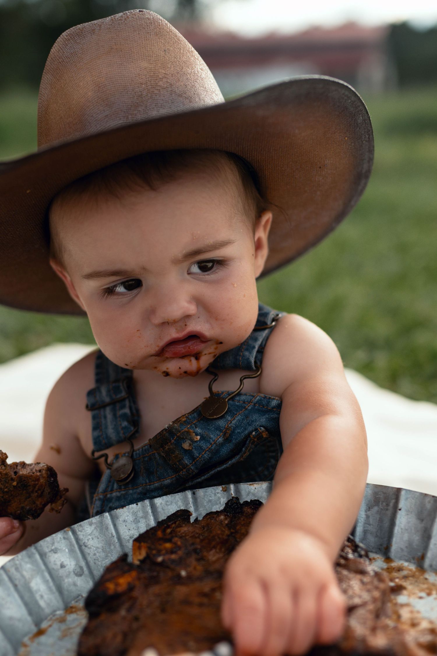 Adorable baby in oversized cowboy hat and denim overalls enjoying a chocolate cake smash outdoors.