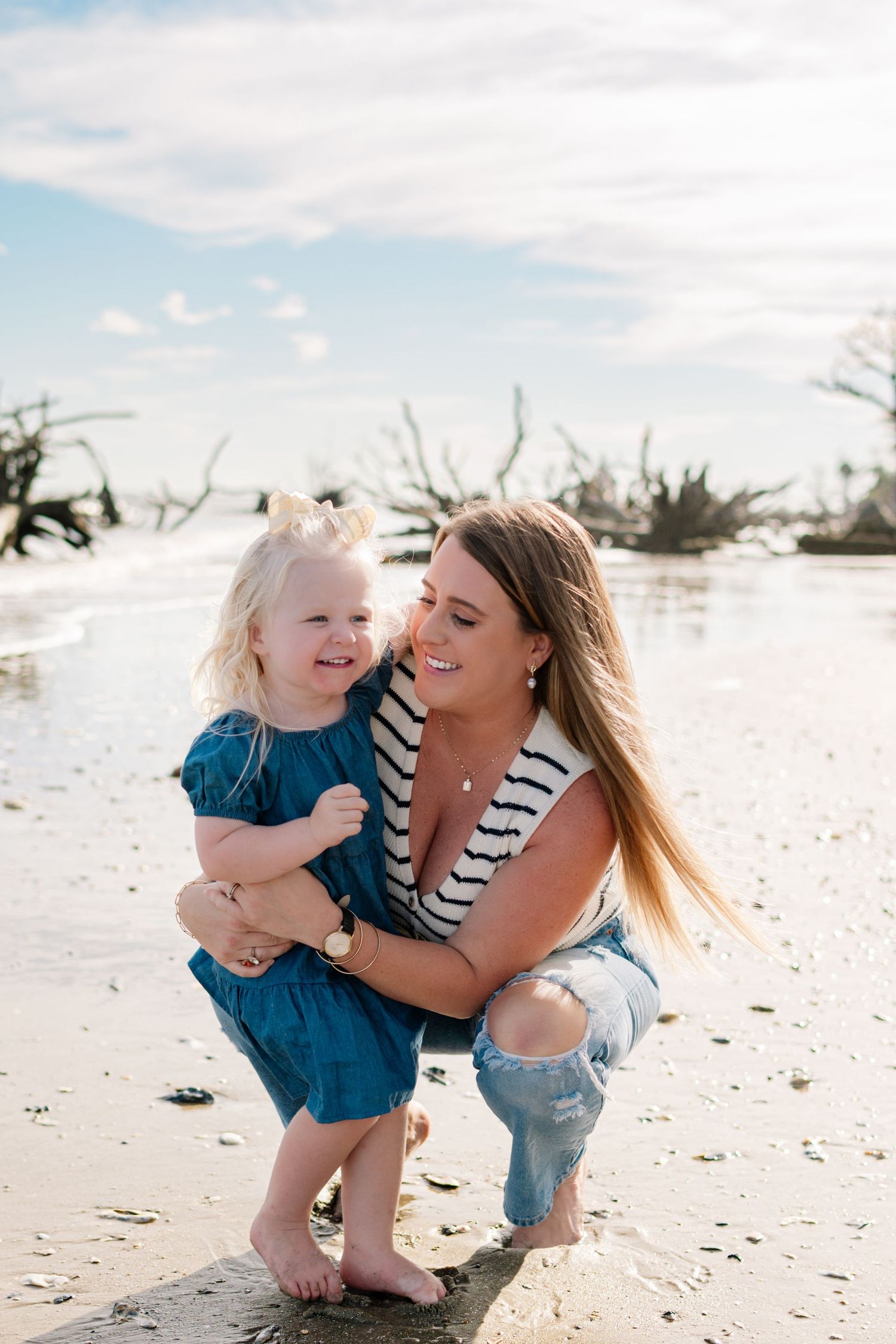 A mother crouches on a beach holding her smiling toddler daughter, both laughing together near the ocean shore.