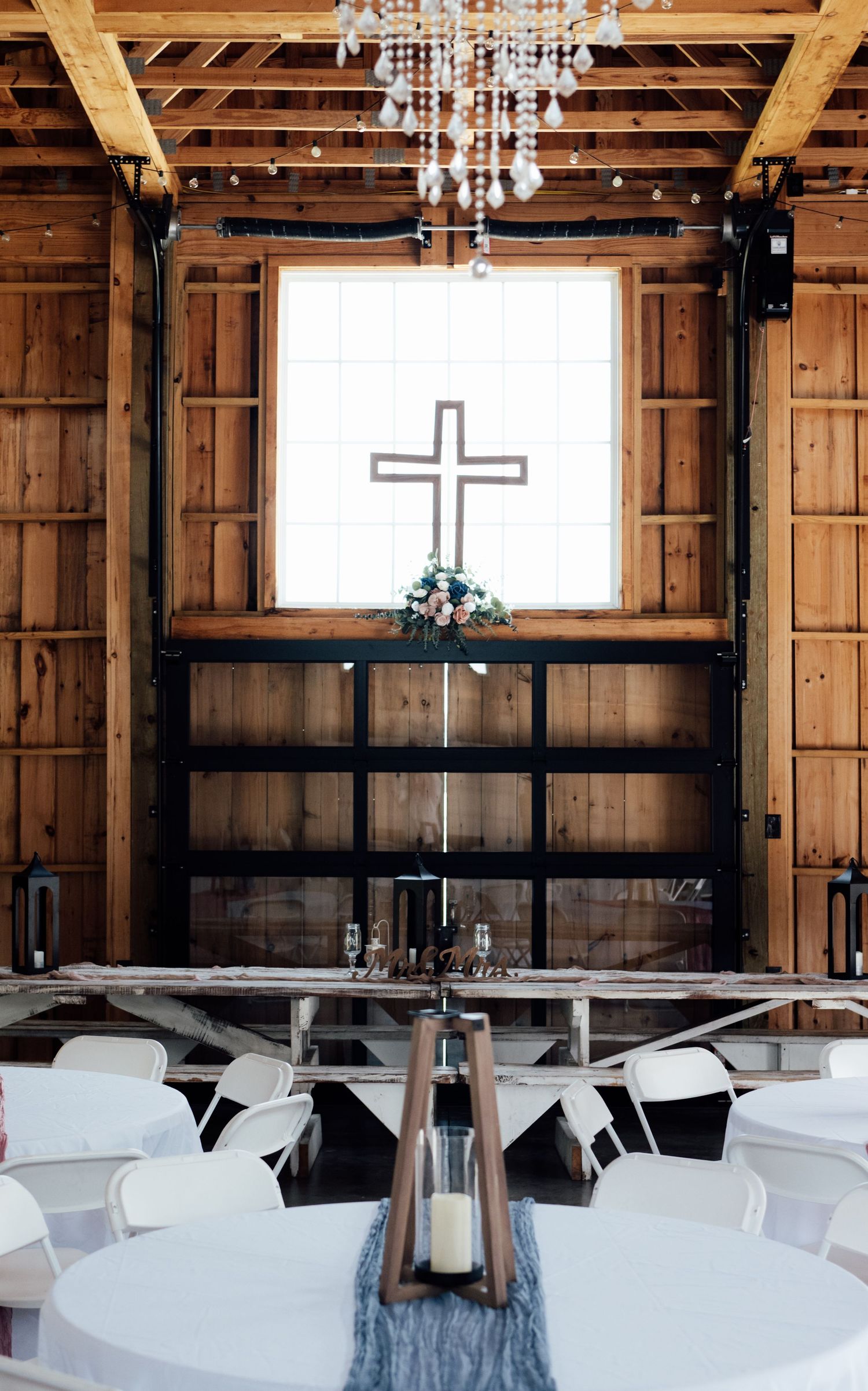 Rustic barn wedding venue with wooden walls, crystal chandelier, cross decor, white chairs, and elegant table settings.