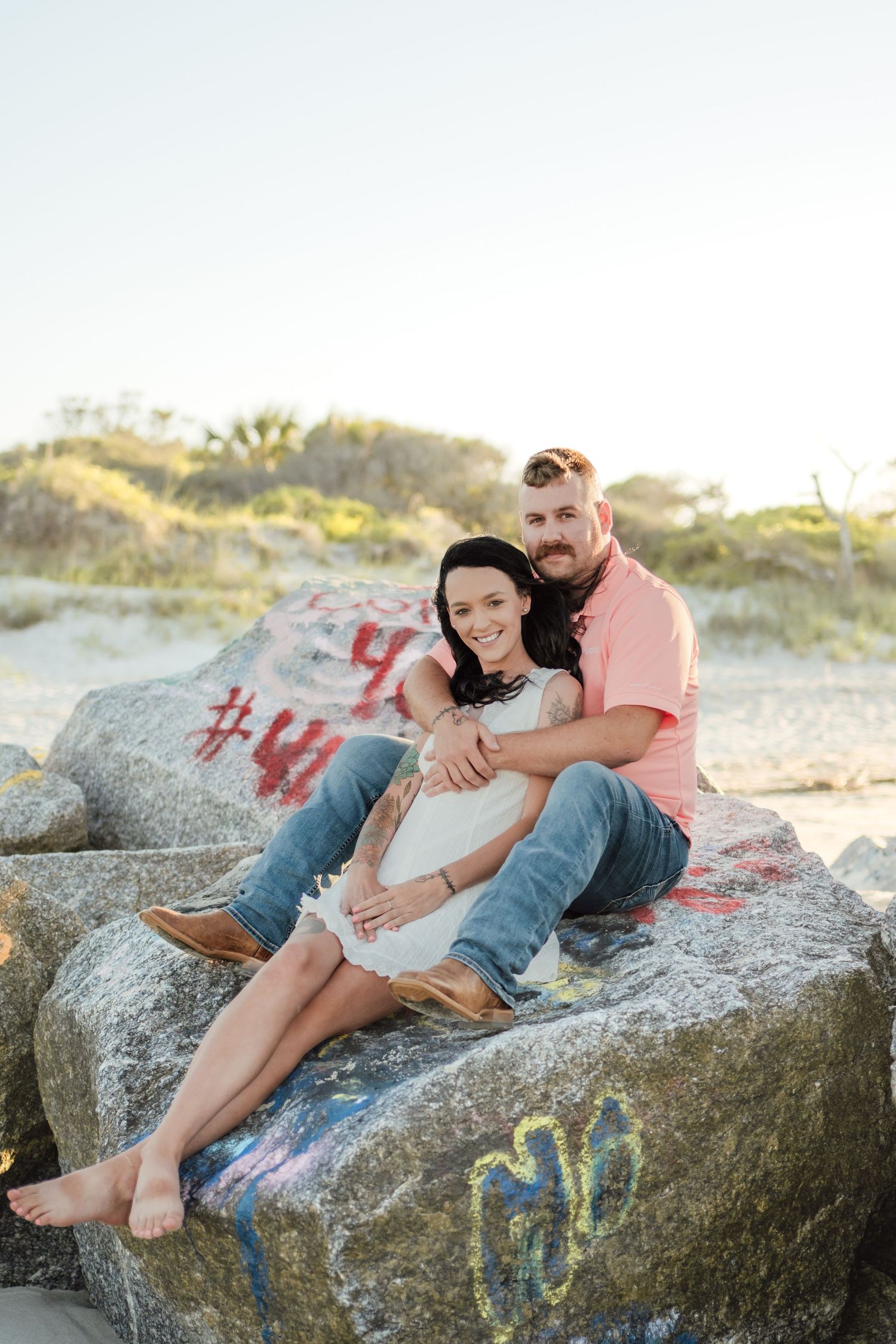 Couple sitting on graffiti-covered rocks at a sandy beach, smiling warmly in casual summer attire.