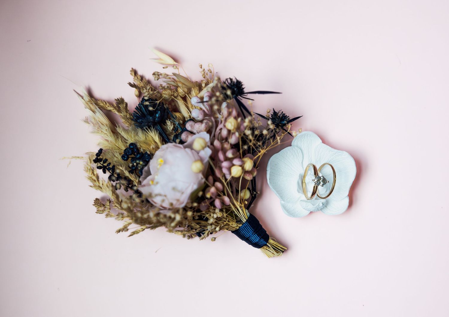 Dried floral boutonniere with purple crystal and wedding rings on blue dish against white background.
