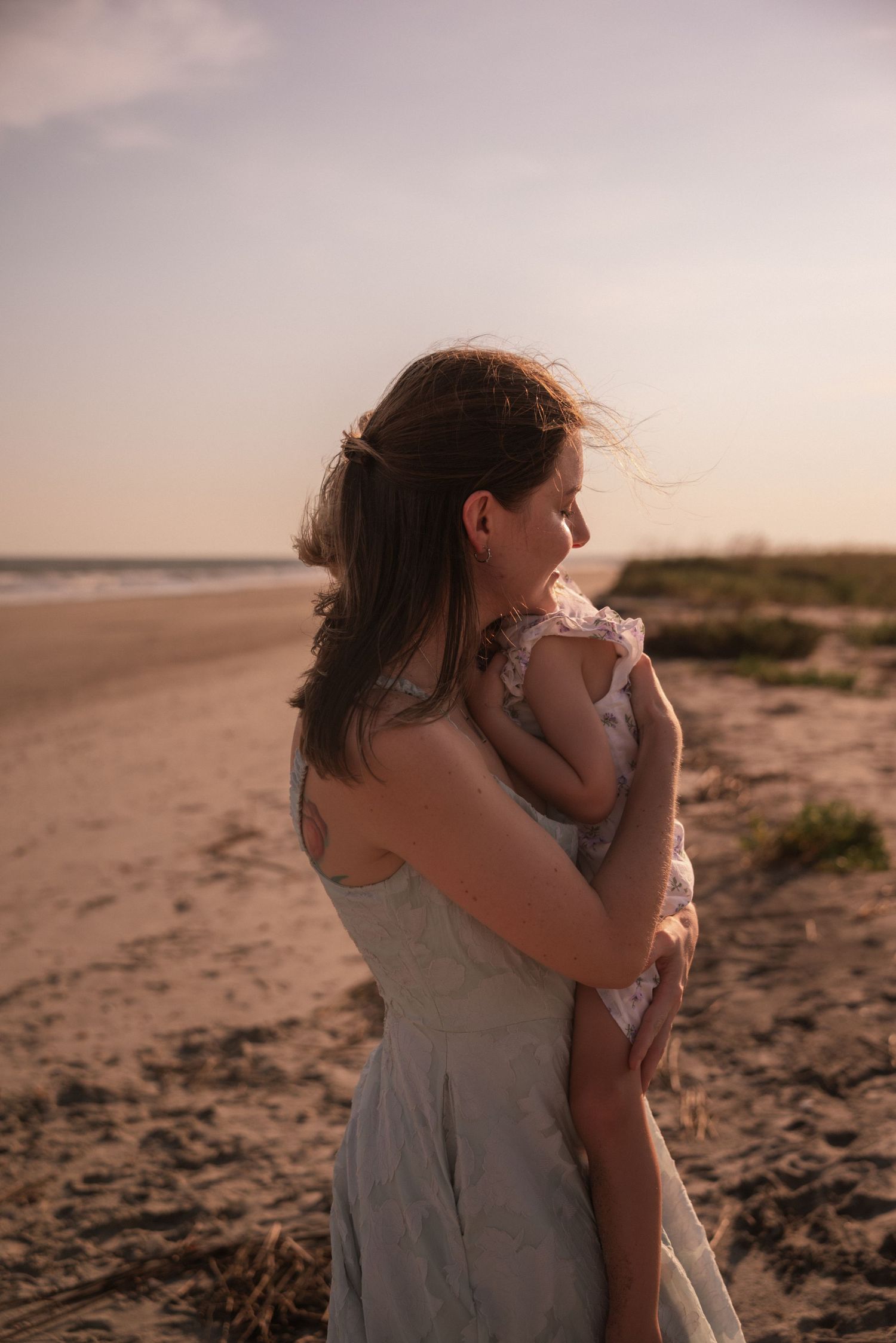 A mother tenderly holds her baby on a sandy beach at sunset, her hair blowing in the breeze.