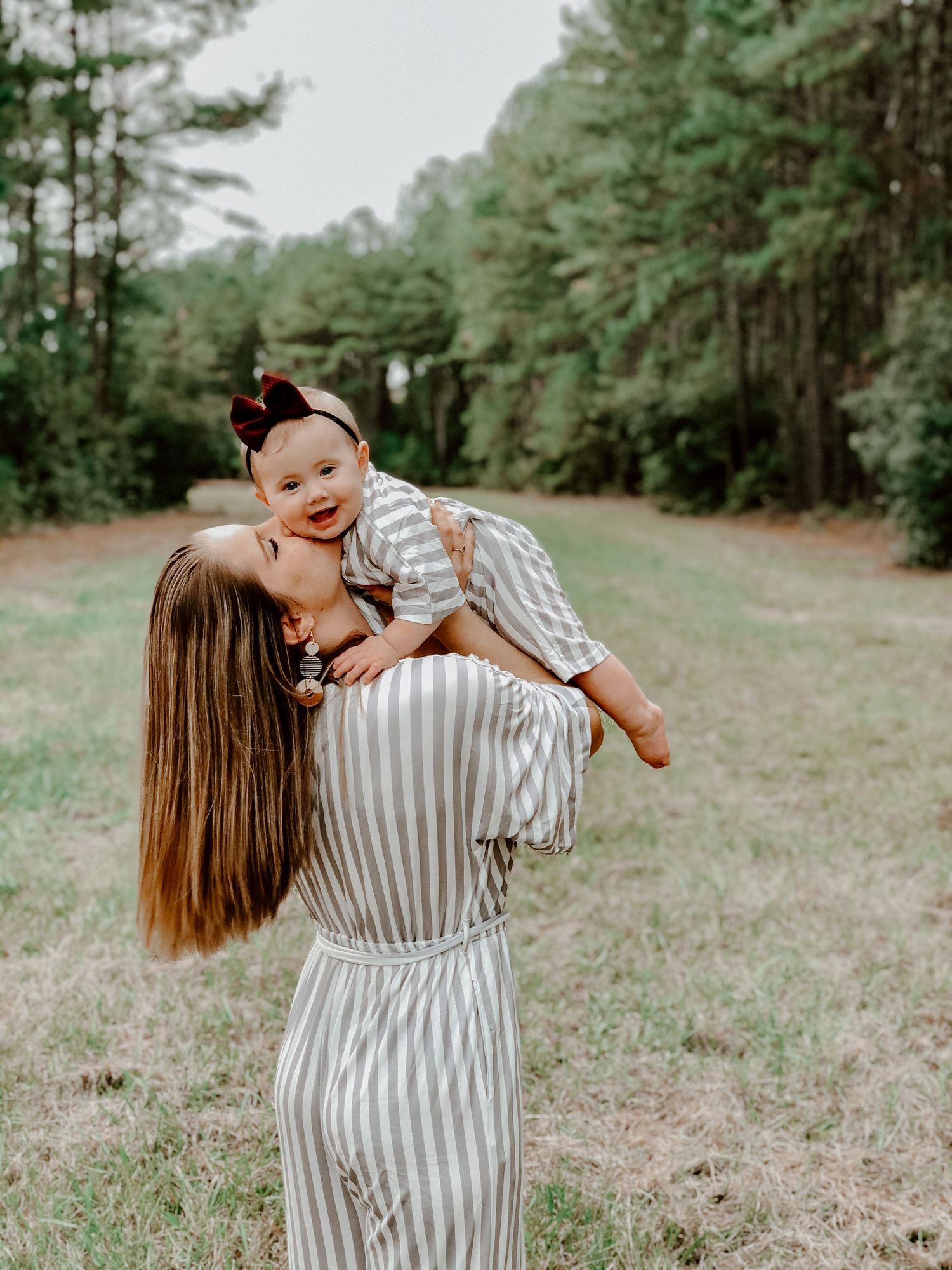A mother in a striped dress kisses her smiling baby girl while lifting her up in a green meadow.