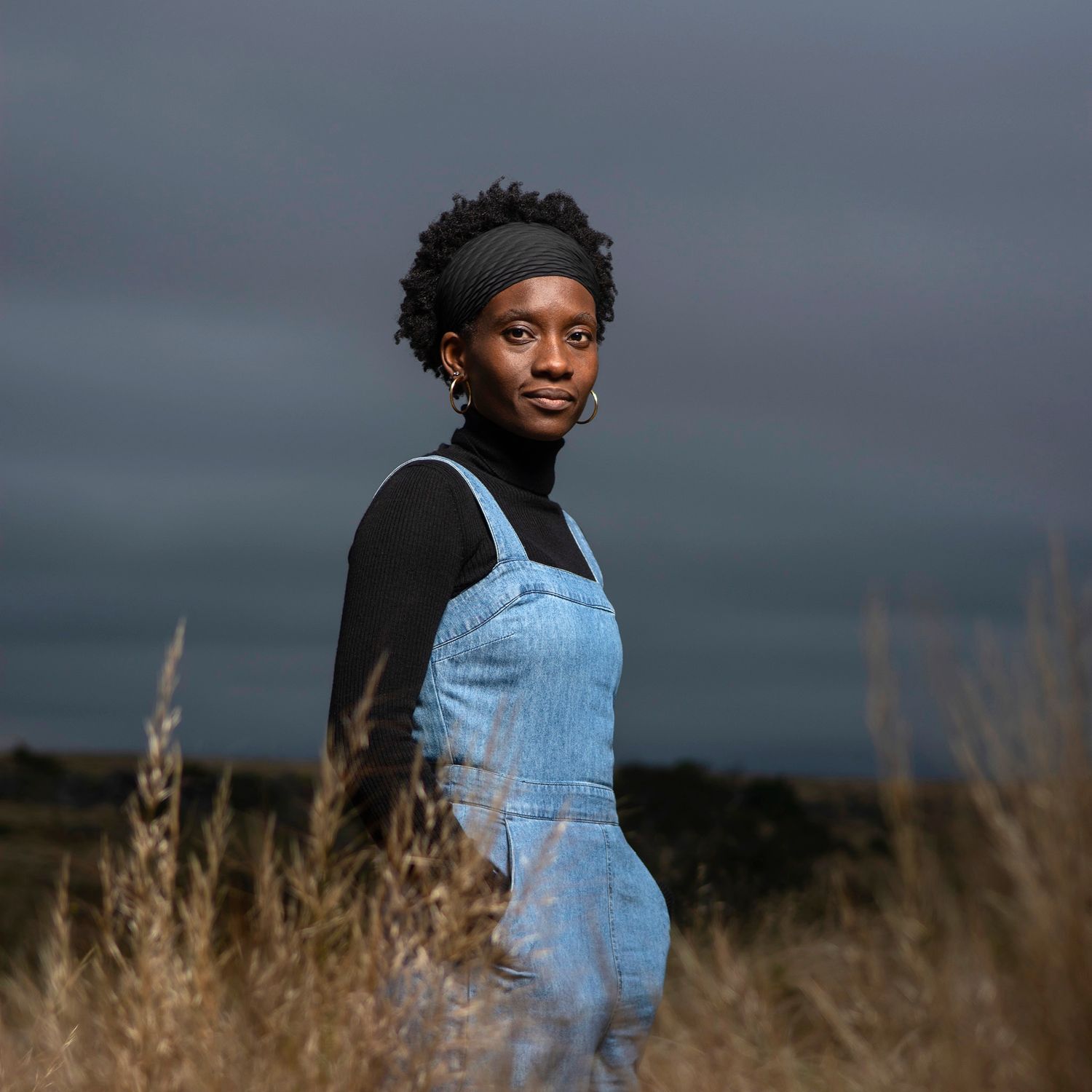 Woman dressed in denim overalls standing behind dry grass.