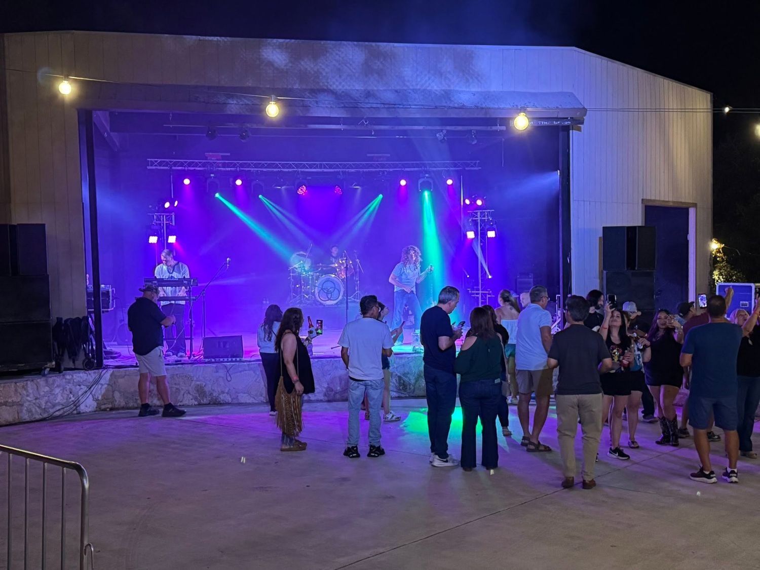 Small crowd watching a band perform on a brightly lit purple and green stage at Josabi's Live nighttime concert in Helotes, Texas.
