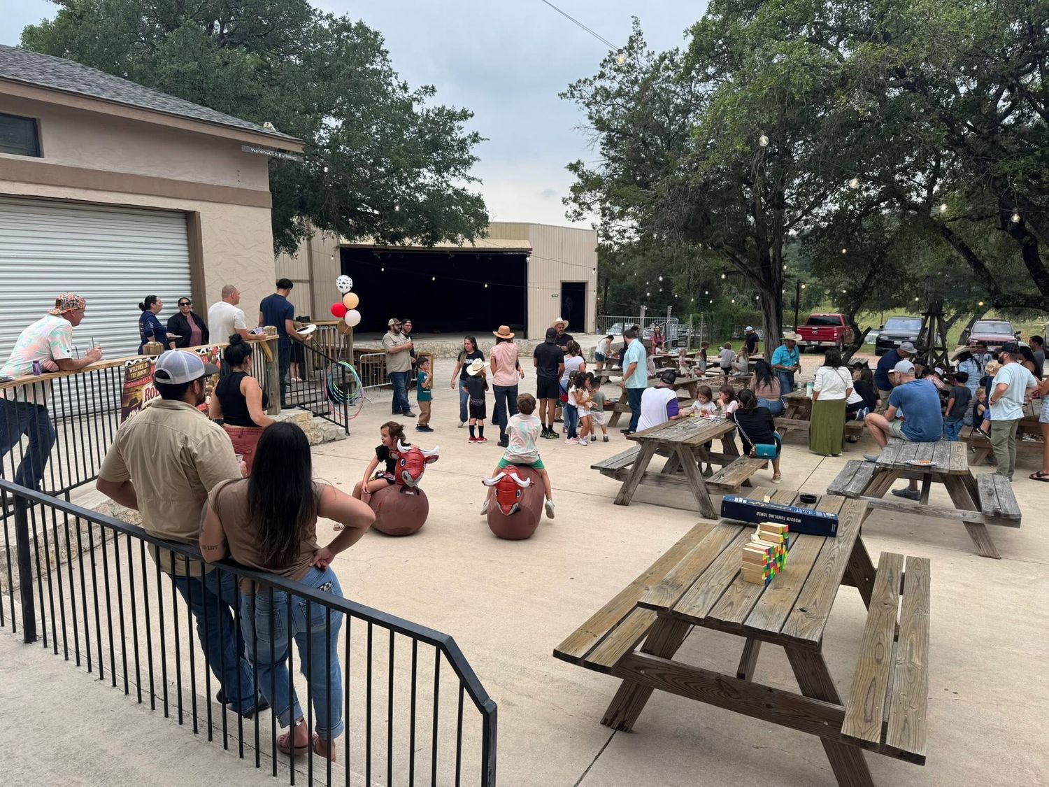 Outdoor event space with picnic tables, families and children gathered near an open-air stage at Josabi's Acres in Helotes, TX.