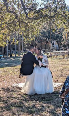Bride and groom share a kiss outdoors under a wooden arch during a sunny wedding ceremony at Josabi's Acres in Helotes, TX.
