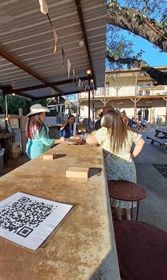 Outdoor bar with menu, women chatting at concrete counter under shaded patio with trees and buildings at Josabi's Acres in Helotes, TX.