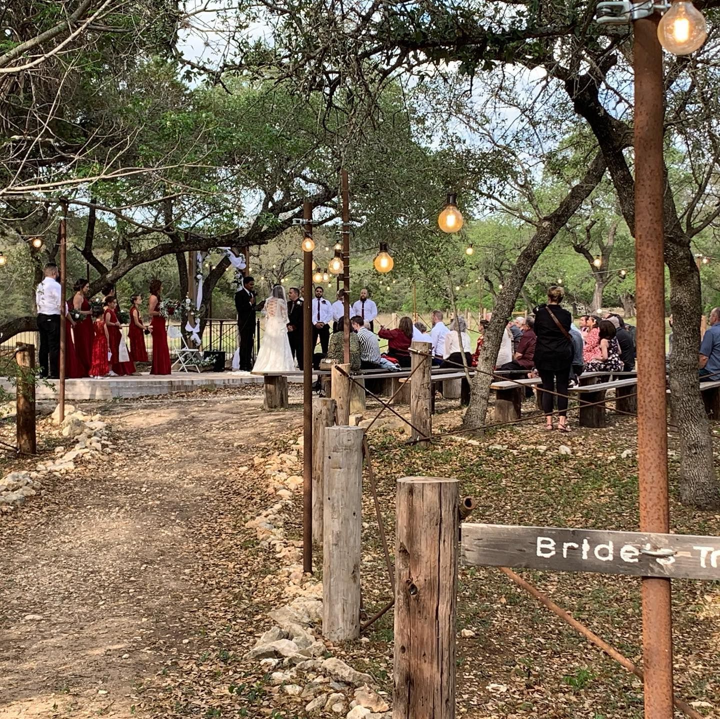 Outdoor rustic wedding ceremony along tree-lined path with red bridesmaid dresses and wooden benches at Josabi's Acres in Helotes, Texas.