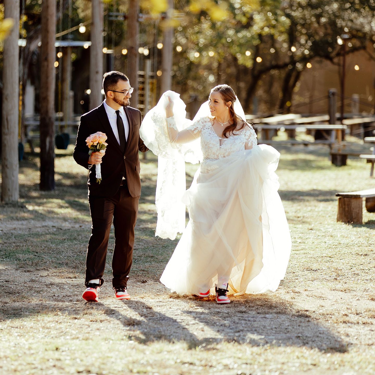 Bride and groom walking outdoors in wedding attire, groom holding bouquet, bride lifting white gown at Josabi's Acres in Helotes Texas.