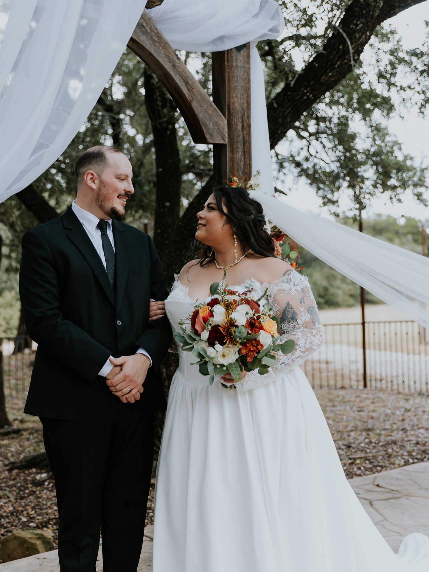 Bride and groom standing under outdoor rustic wooden arch, white draping, bride holding colorful bouquet at Josabi's Acres in Helotes TX.