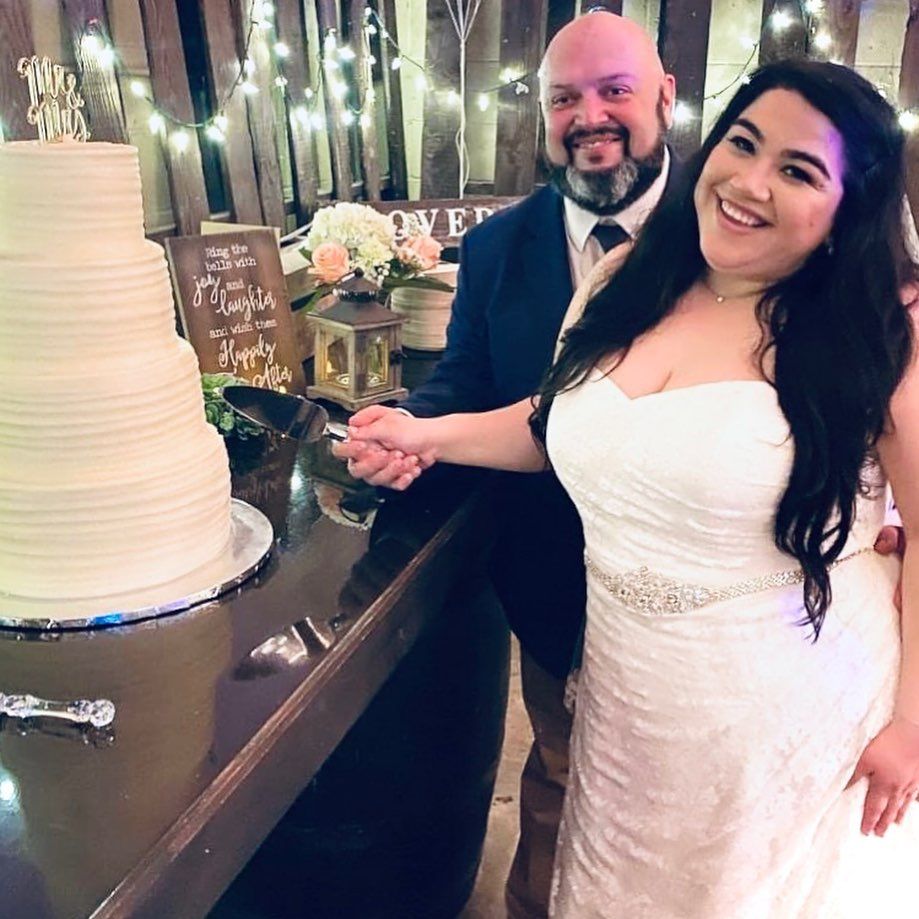 Bride and groom cutting elegant white tiered wedding cake in rustic barn venue with string lights at Josabi's Acres in Helotes, Texas.