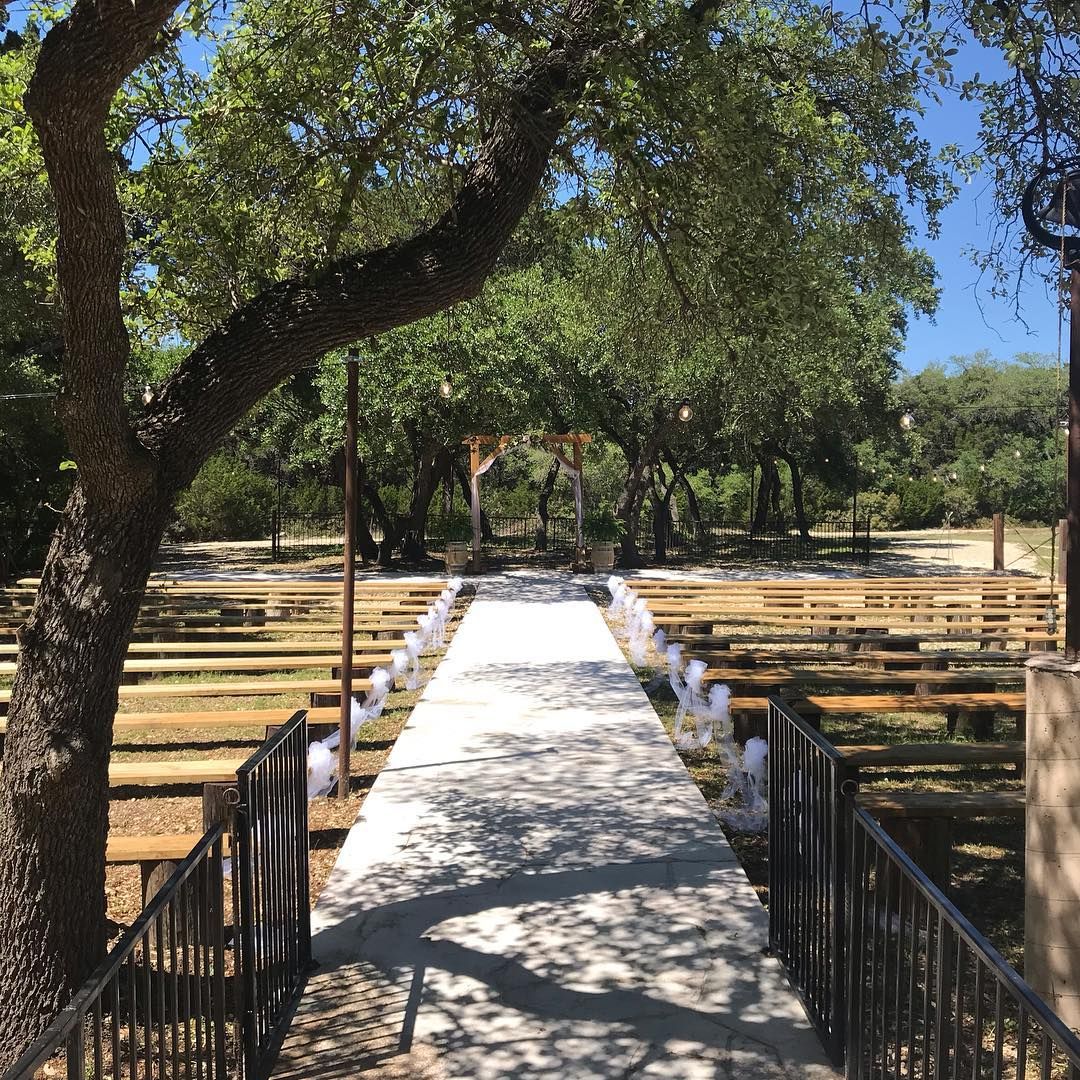 Sunny outdoor wedding aisle with white runner, wooden pews, floral decorations, and a rustic wooden arch at Josabi's Acres in Helotes, Texas
