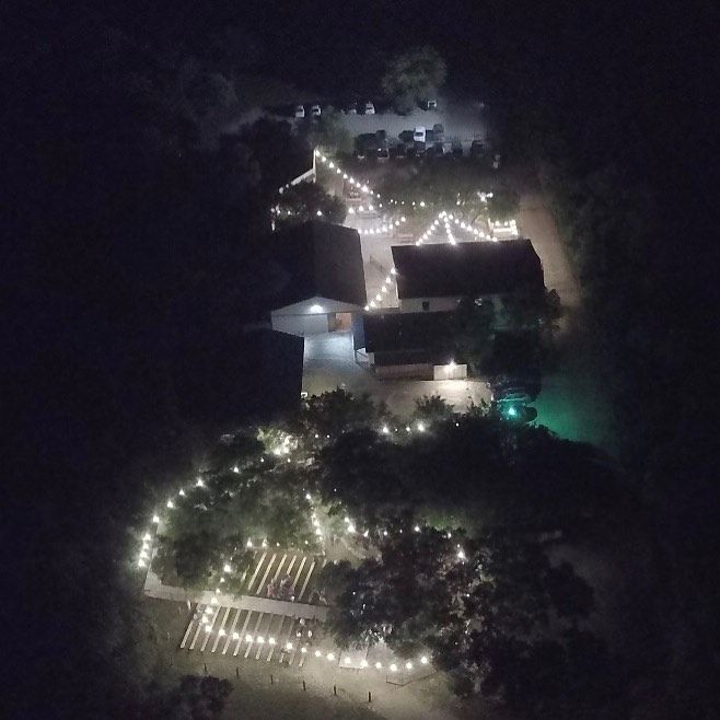 Aerial night view of a rustic private event venue illuminated by string lights surrounded by trees at Josabi's Acres in Helotes, Texas