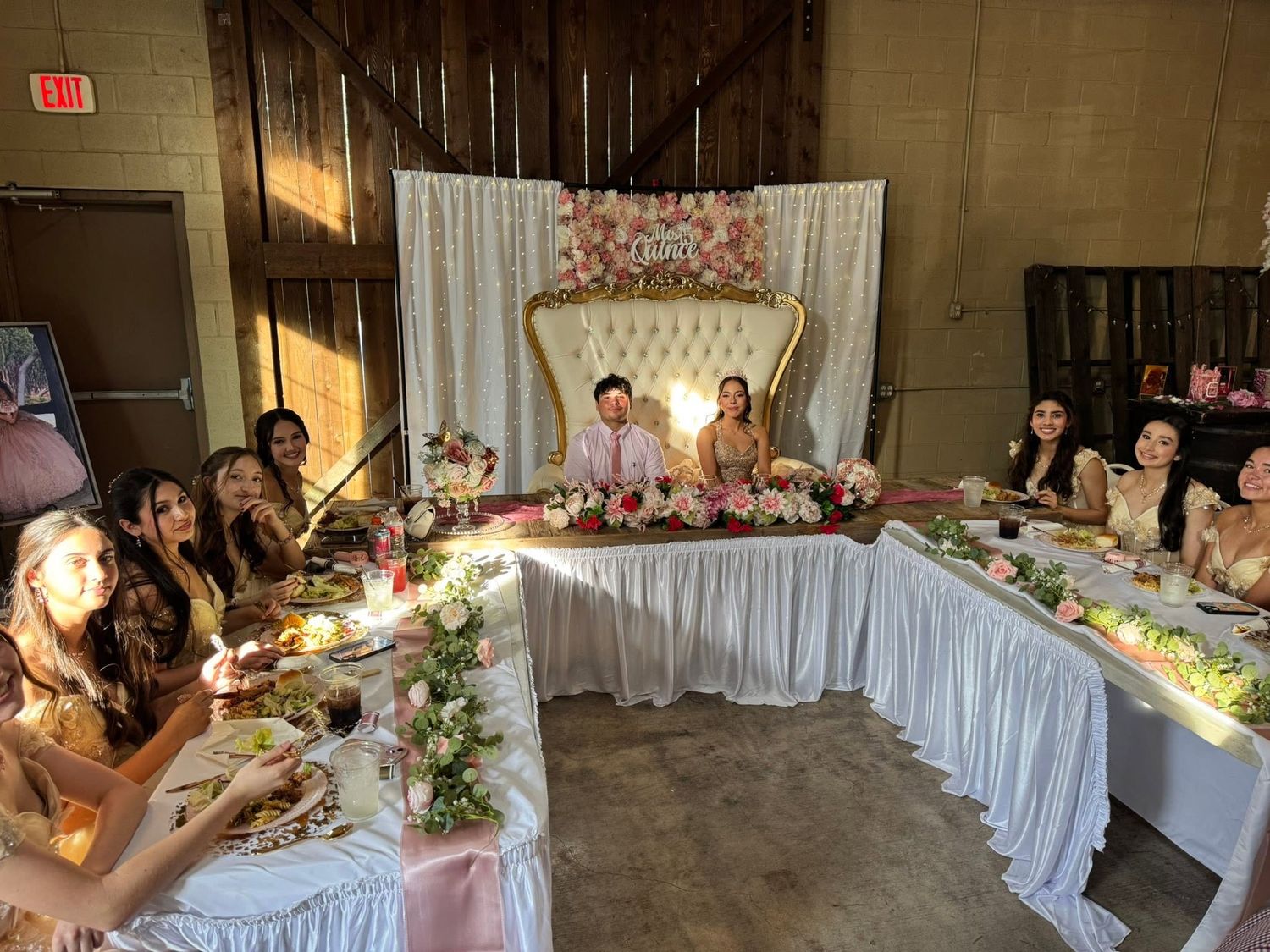 Quinceañera court seated at a decorated table with floral arrangements in a barn venue at Josabi's Acres in Helotes, TX.