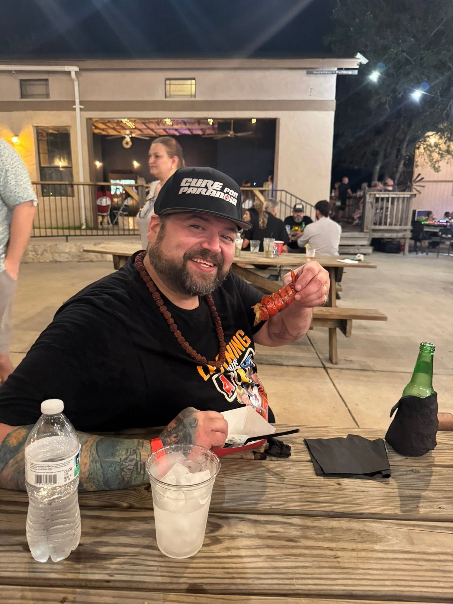 Bearded man in black t-shirt and cap holds crawfish at outdoor picnic table with drinks at Josabi's Acres in Helotes, TX.