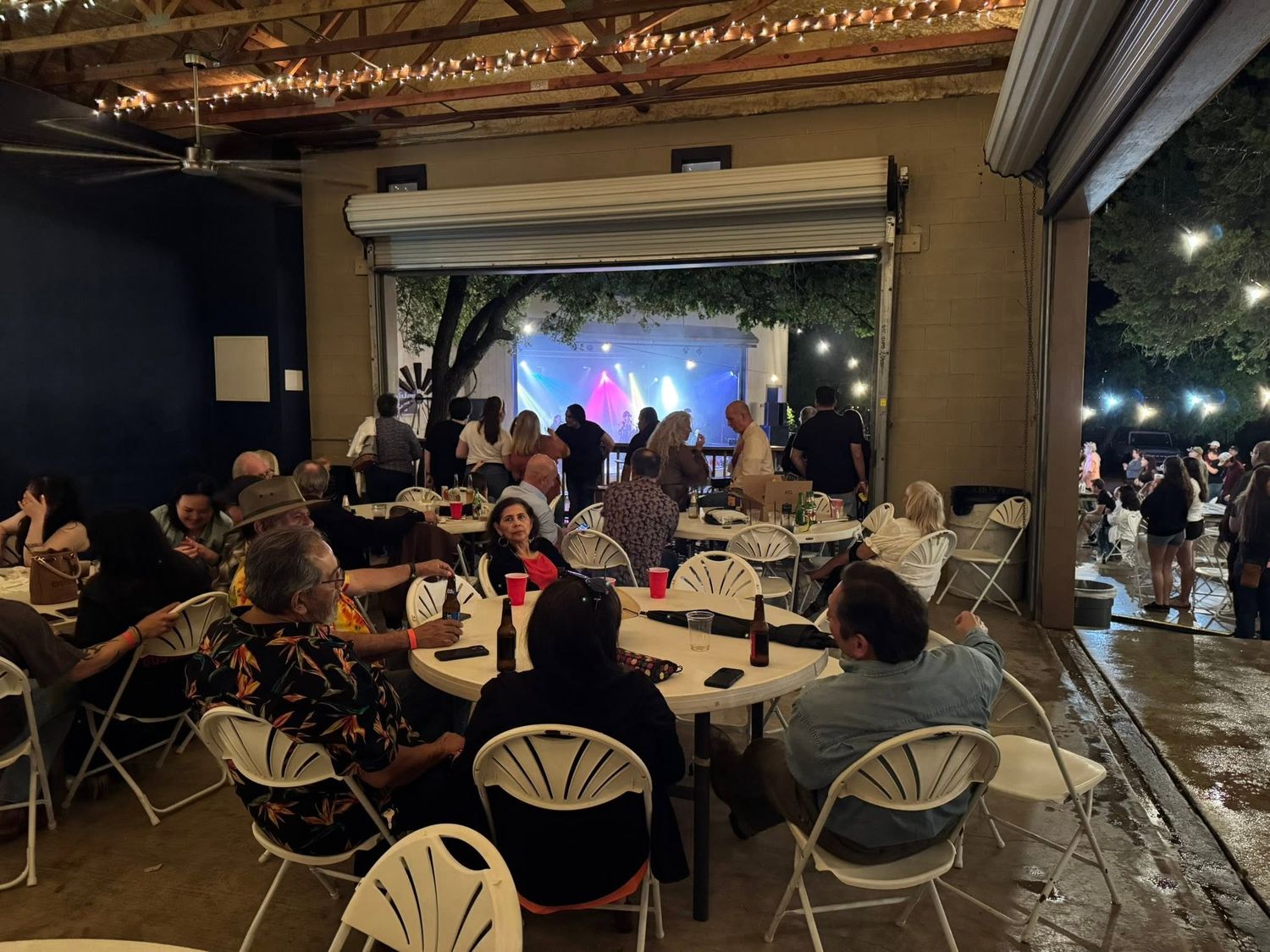 Indoor venue round tables and guests seated watching a performer on a lit stage through an open rollup door at Josabi's Acres in Helotes TX.