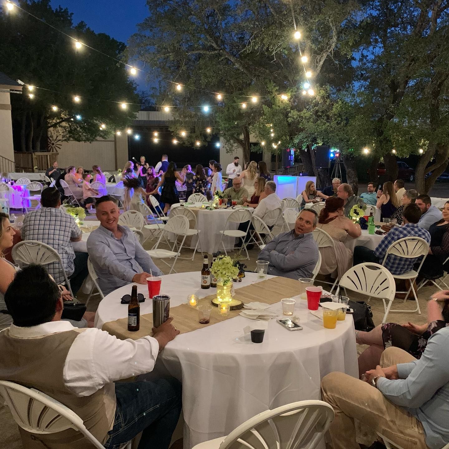 Wedding reception guests seated at round tables under string lights with dance floor glowing in background at Josaib's Acres in Helotes TX.