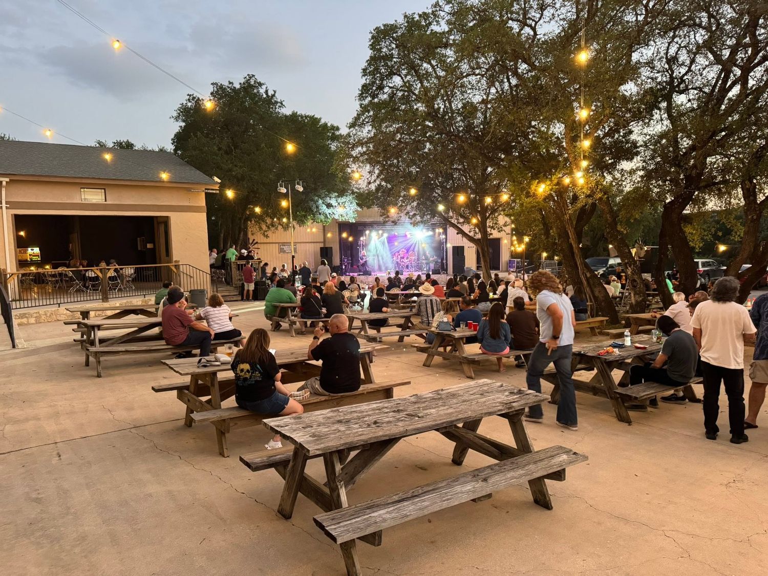 Outdoor concert venue picnic tables, string lights, large crowd near a colorfully lit performance stage at Josabi's Acres in Helotes TX.