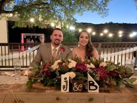 Smiling couple at wedding sweetheart table decorated with burgundy floral centerpiece and monogram J&B sign at Josabi's Acres in Helotes, TX