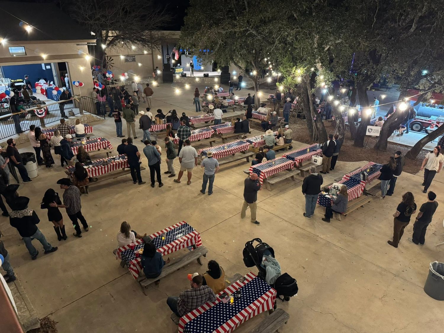 Aerial view outdoor evening event patriotic tablecloths, string lights, groups of people mingling on patio at Josabi's Acres in Helotes TX.