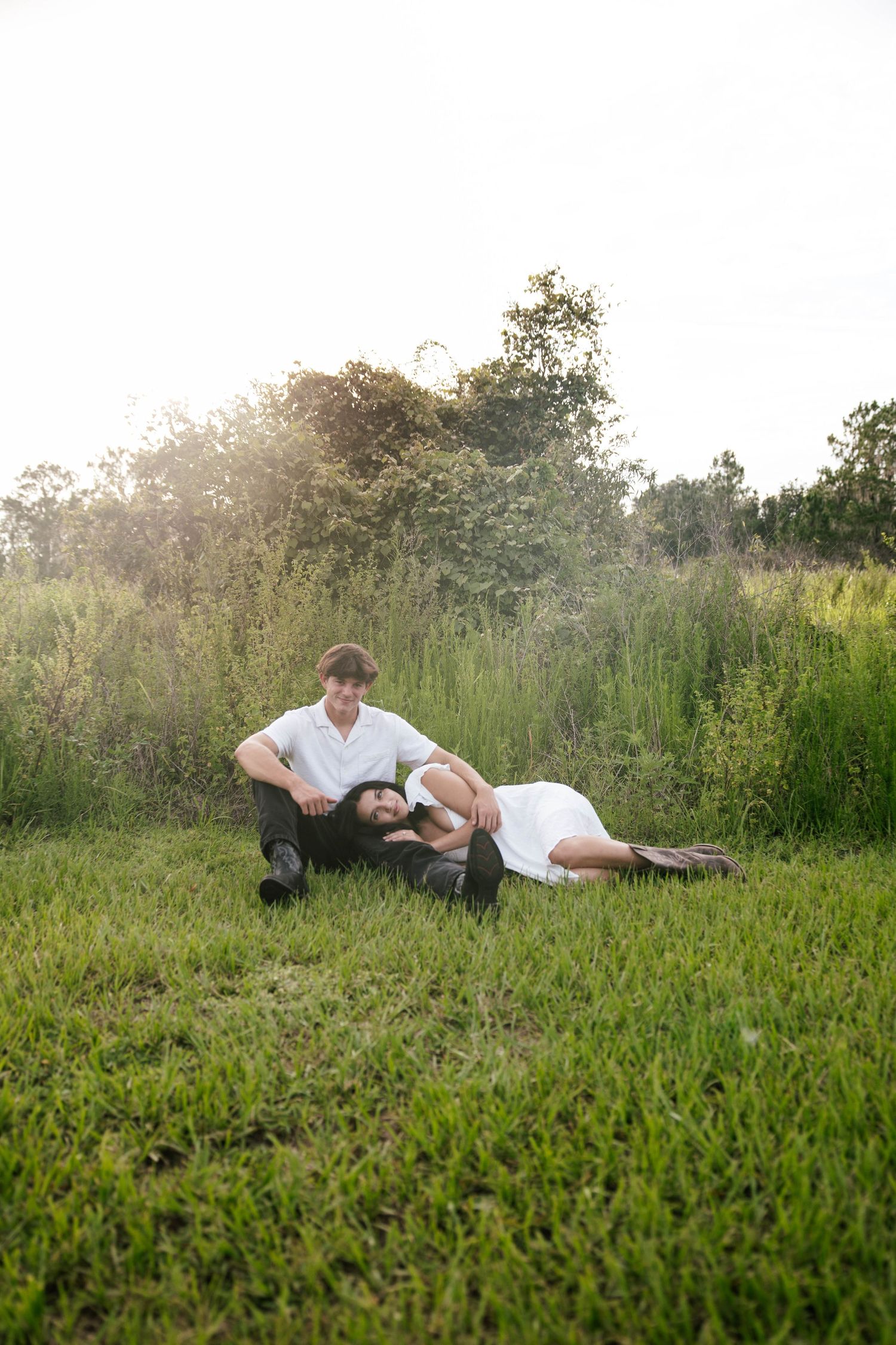 Couple relaxing on green grass field, woman in white dress lying on man's lap, surrounded by lush trees in golden sunlight.
