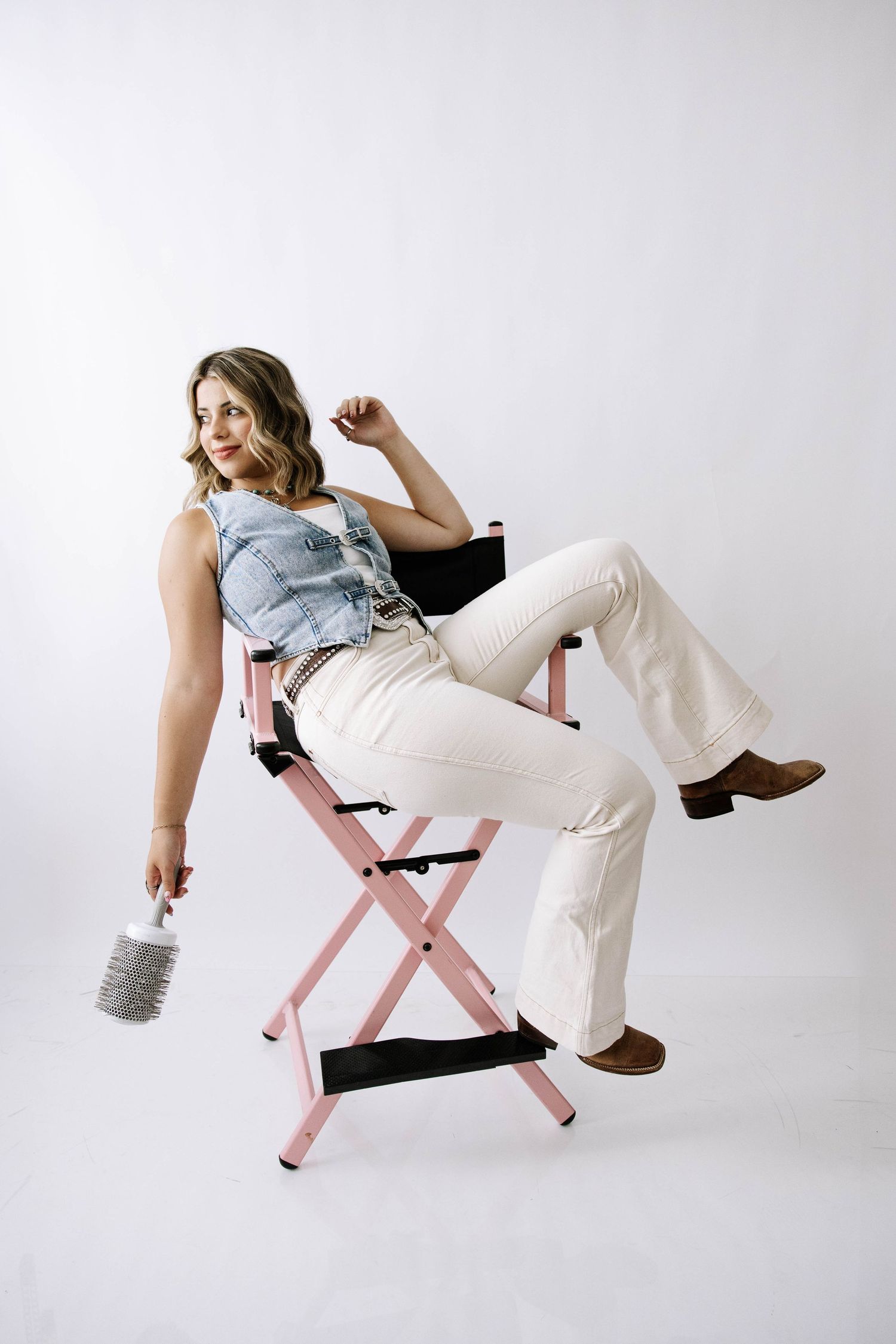 Woman in denim vest and cream flare pants posing playfully on pink directors chair.