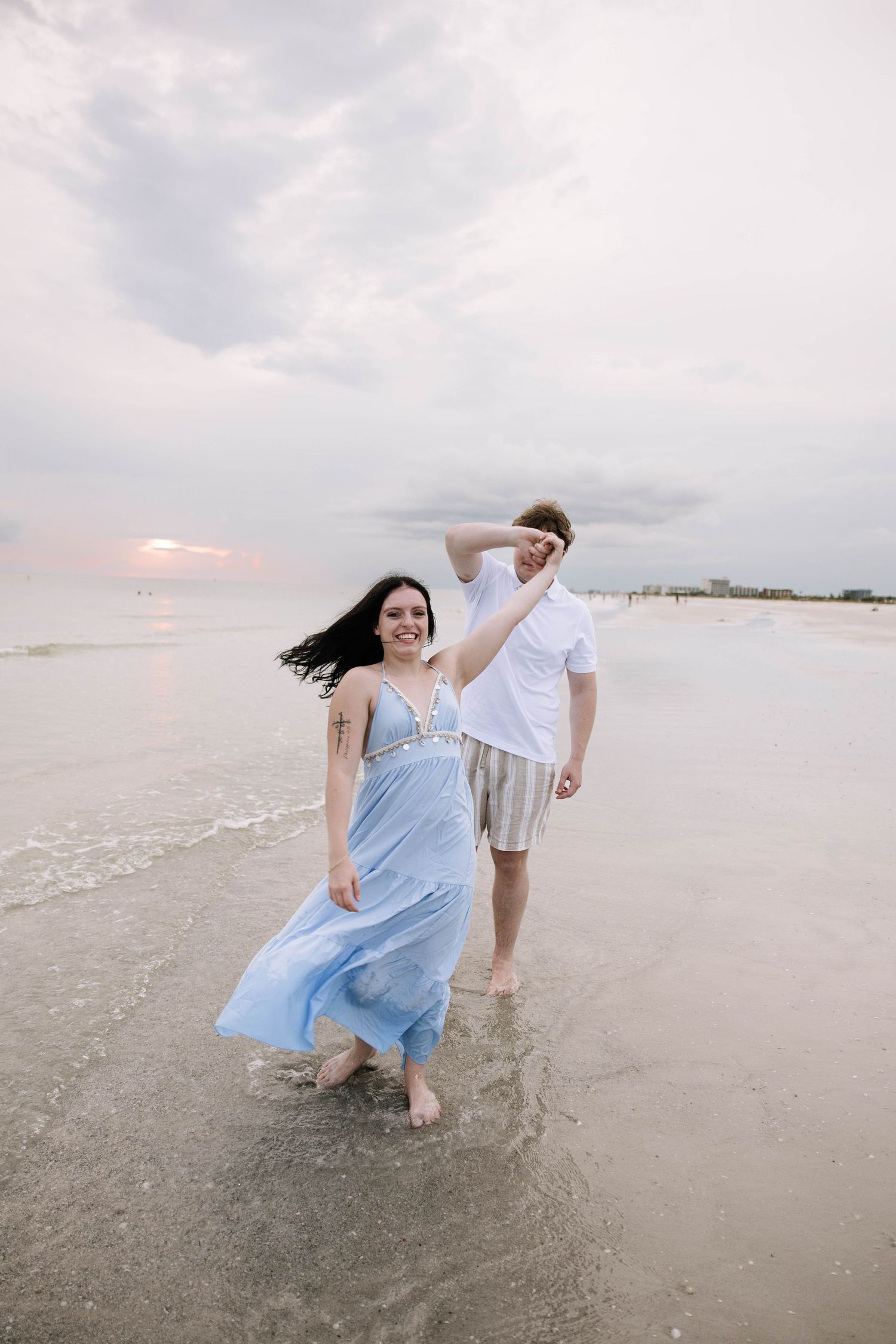 Couple walking barefoot on beach at sunset, woman in flowing blue dress holding man's hand in white shirt and striped shorts.