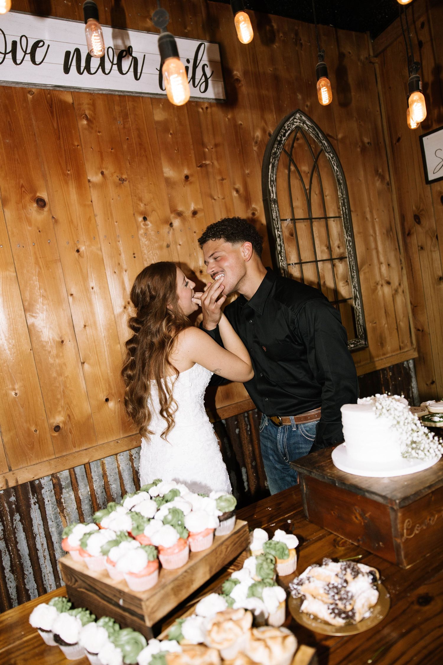 Bride and groom sharing a sweet moment at rustic wedding dessert table with cupcakes and white cake.
