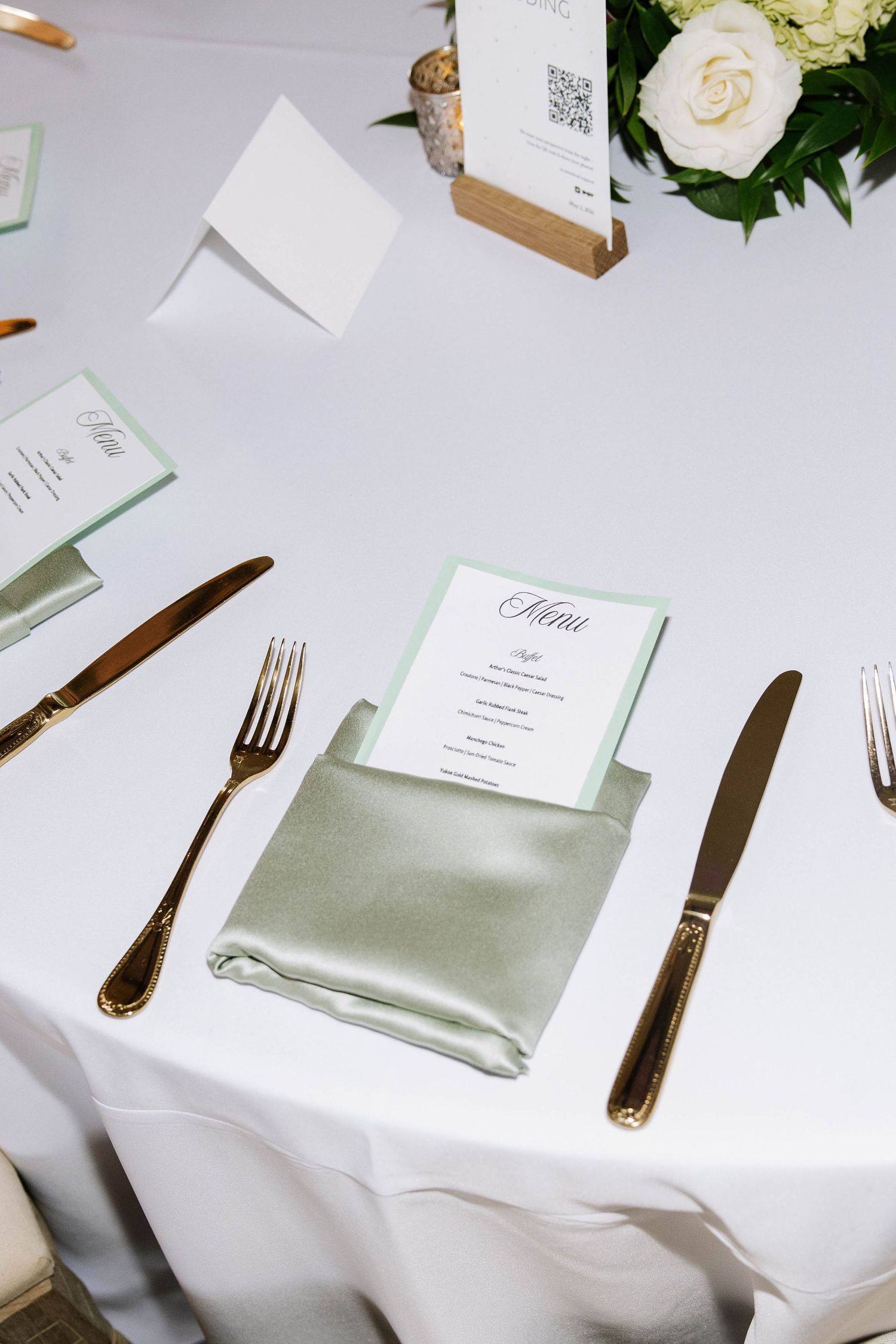 Elegant wedding reception table setting with sage green napkin, gold cutlery, menu card, and white roses.