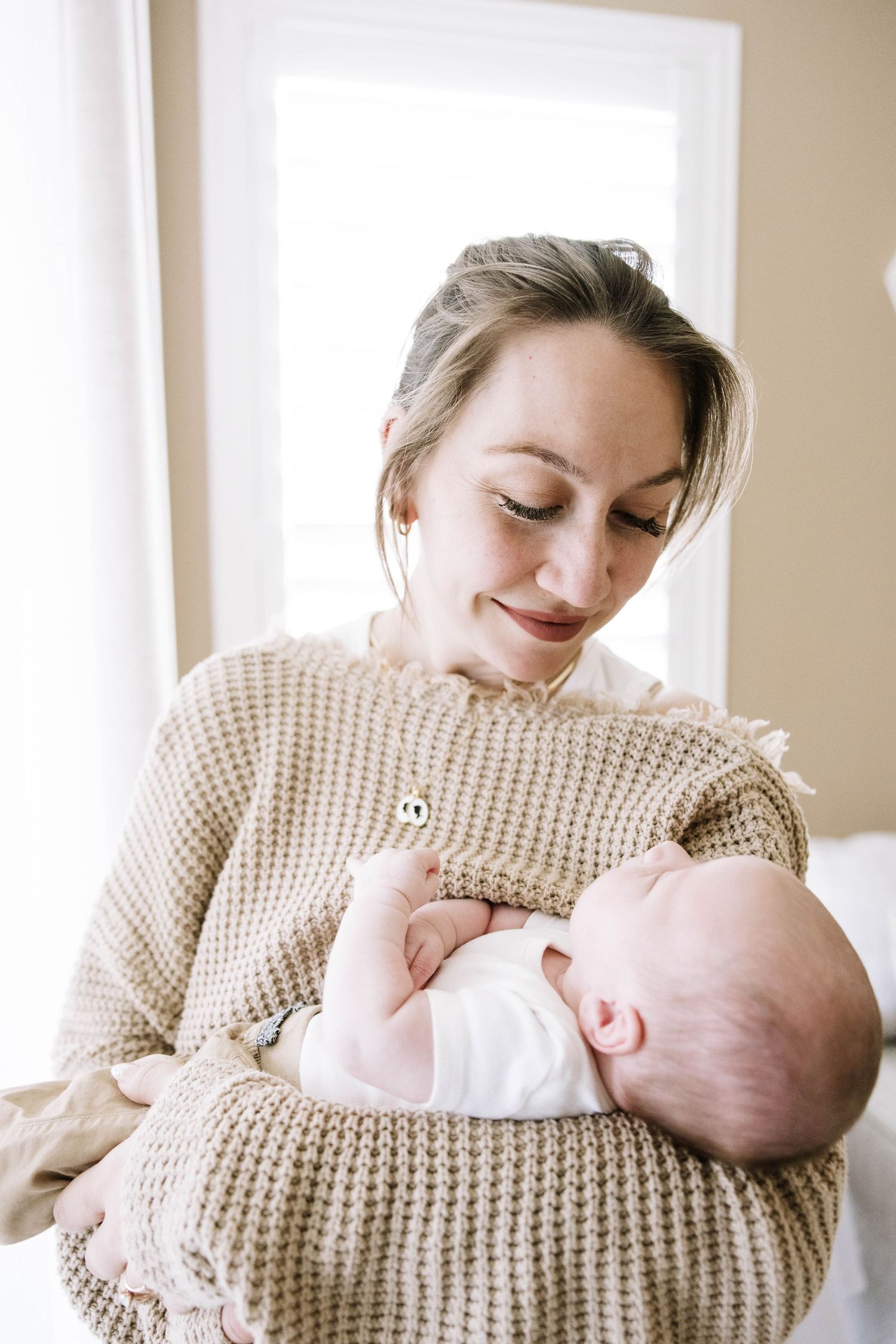 Smiling mother in beige waffle knit sweater tenderly holding her newborn baby near a bright window. Home in Lakeland Florida.