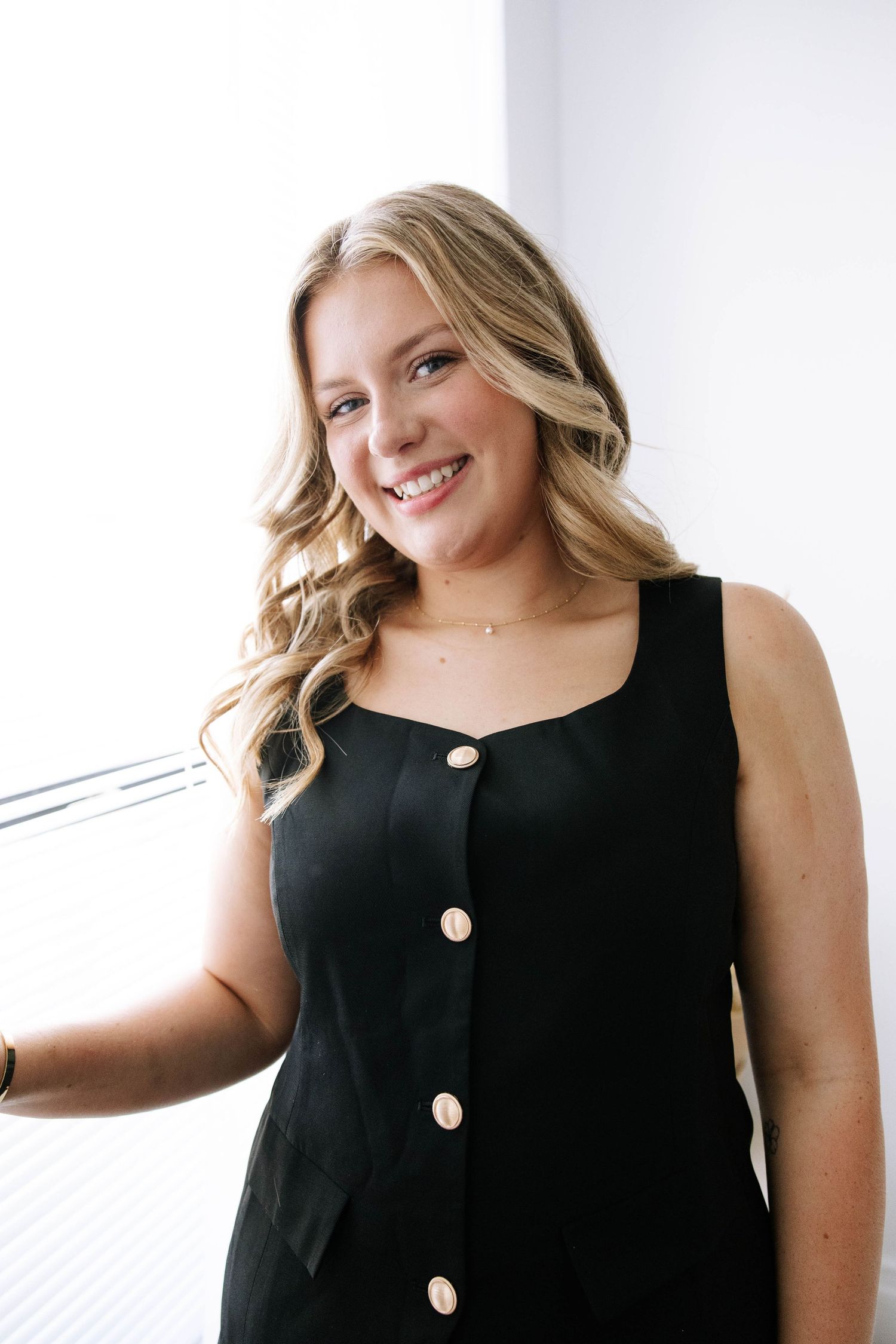 Smiling blonde woman in black sleeveless button dress poses near bright window. Bartow Florida