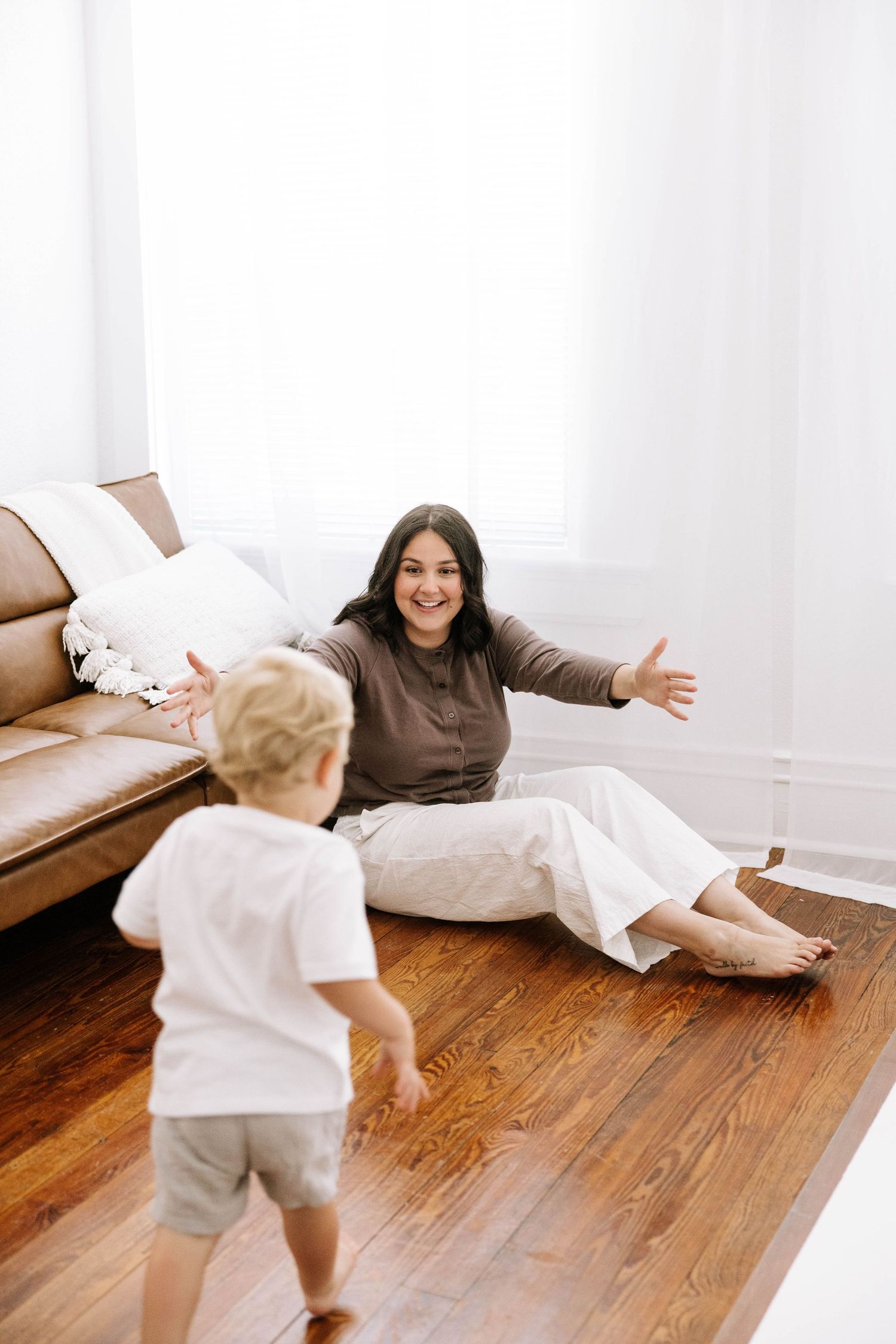 Happy mother sitting on hardwood floor with arms open welcoming her toddler.