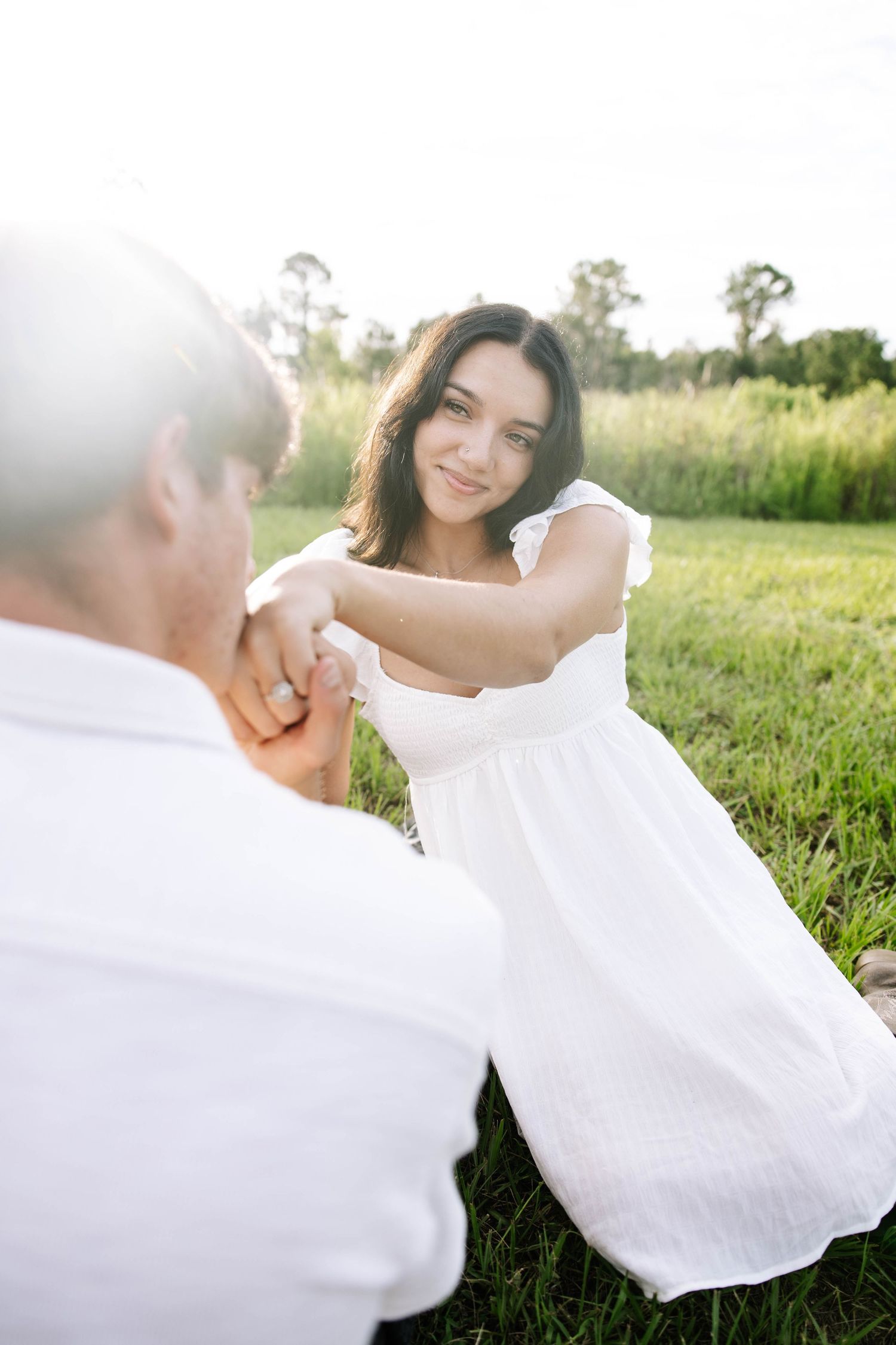 Young woman in white dress holds hands with man in sunlit green field, smiling warmly in romantic outdoor setting.