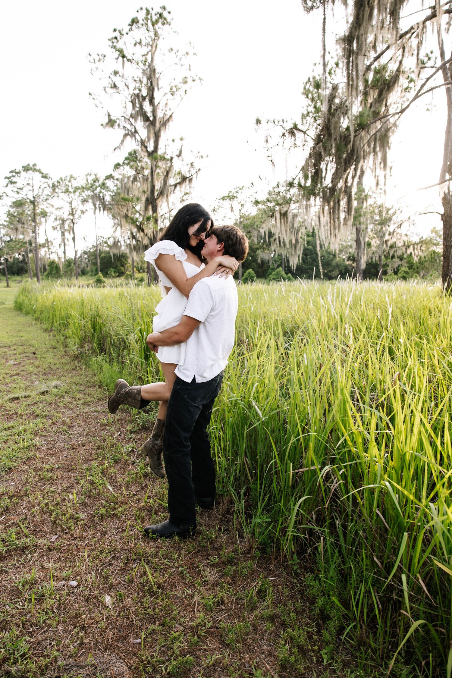 Couple embracing and kissing in a lush green field with Spanish moss-draped trees in a romantic outdoor setting.