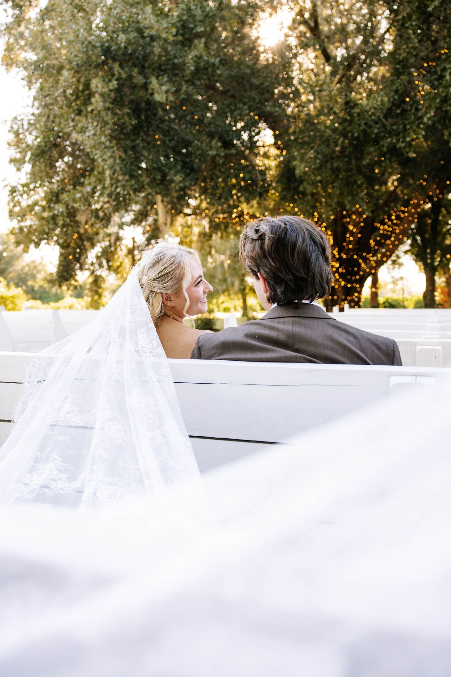 Bride and groom sit together on white bench, bride's lace veil flowing, surrounded by trees with fairy lights at outdoor wedding.