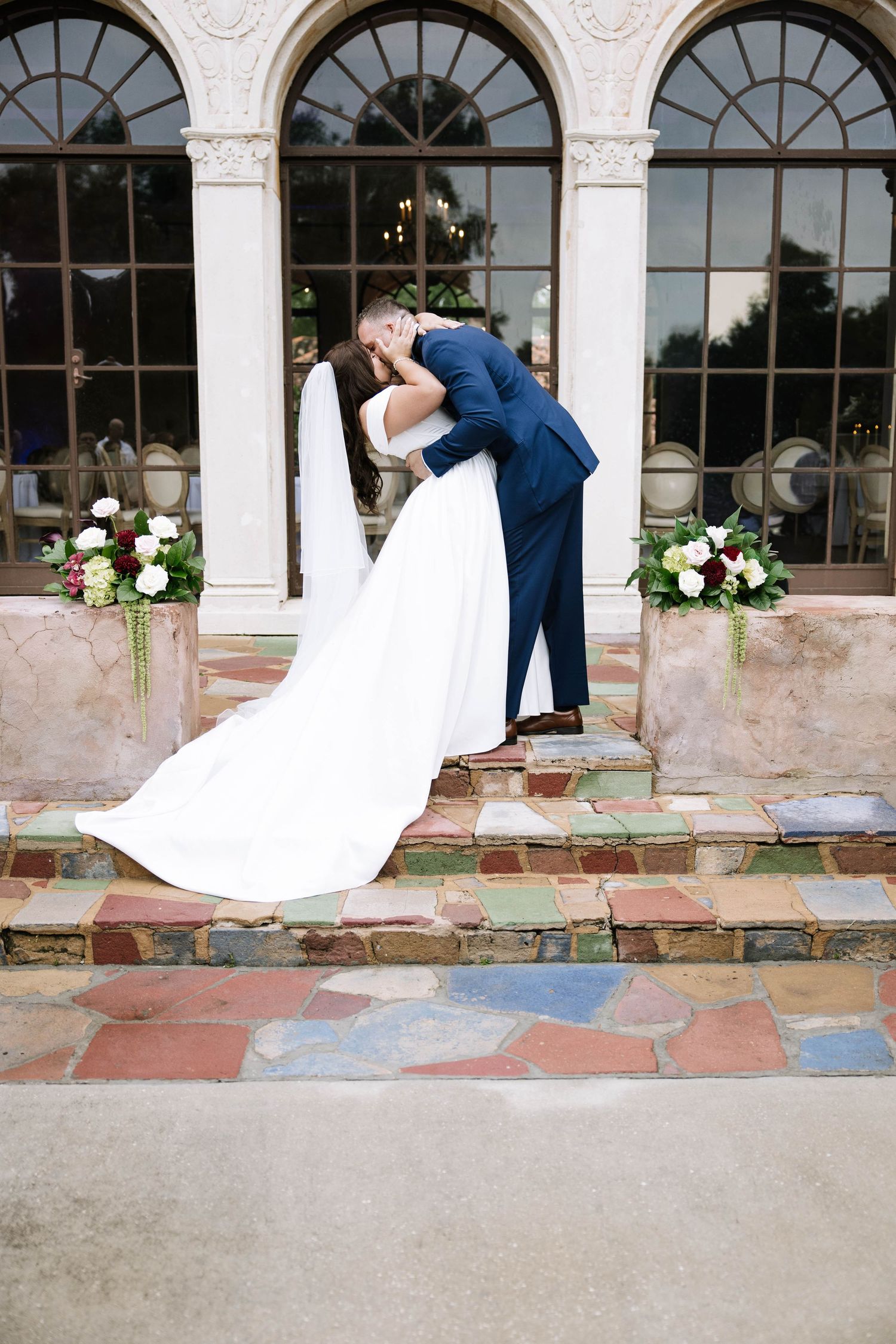 Bride and groom sharing a romantic dip kiss on colorful stone steps outside an elegant venue with floral arrangements.
